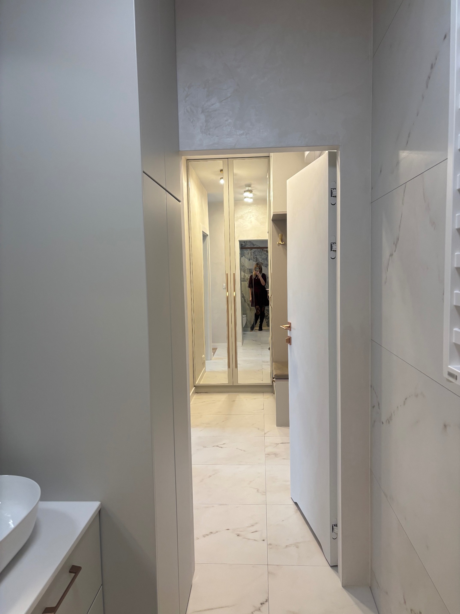 Nowoczesny przedpokój z szafą na wymiar z lustrzanymi drzwiami, marmurowa podłoga i ściany w odcieniach szarości. Widoczna osoba robiąca zdjęcie w lustrze.