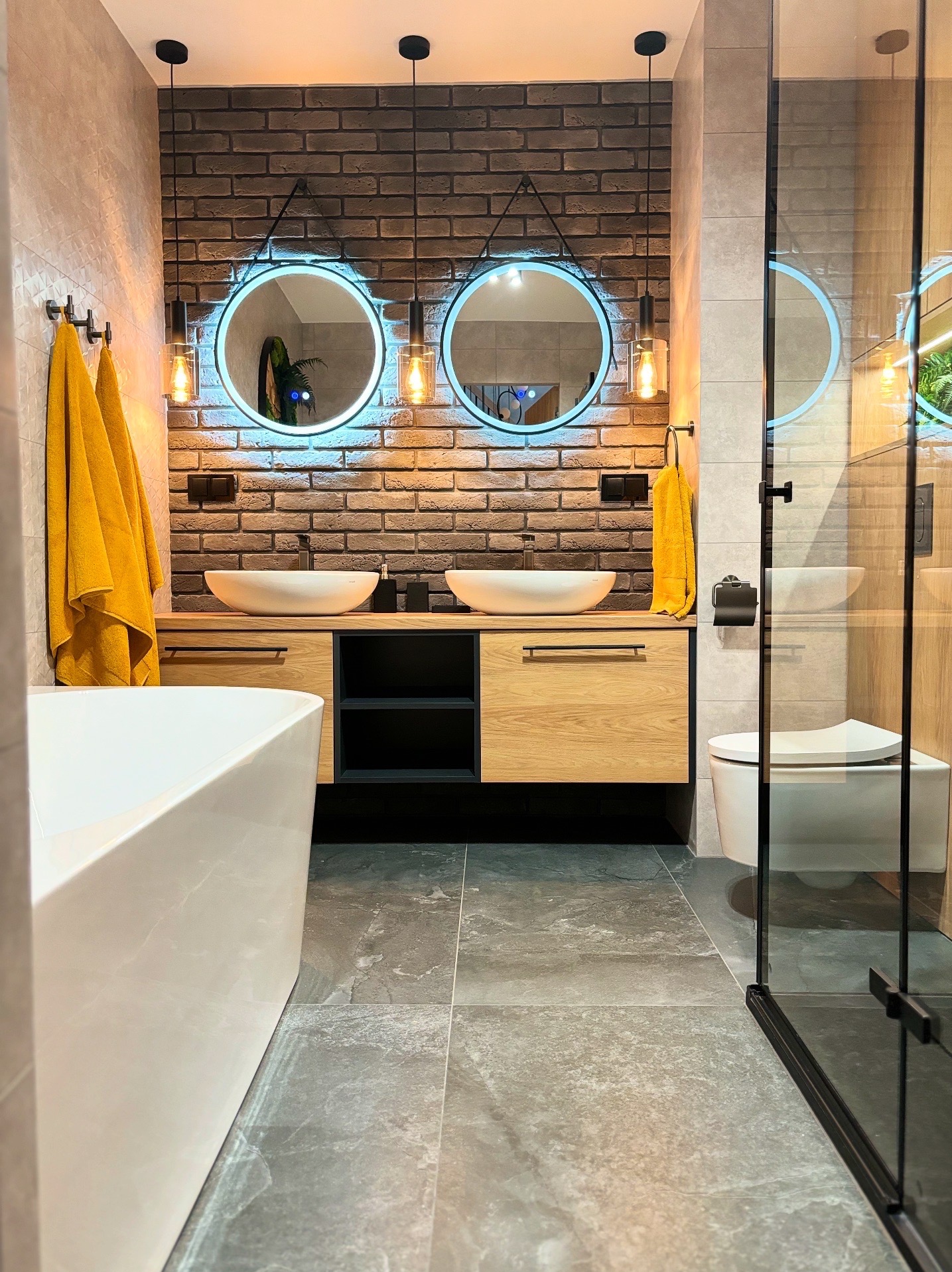 Nowoczesna łazienka z ceglaną ścianą, okrągłymi lustrami z podświetleniem LED, drewnianą szafką pod umywalki i szarą podłogą. Żółte ręczniki dodają koloru.