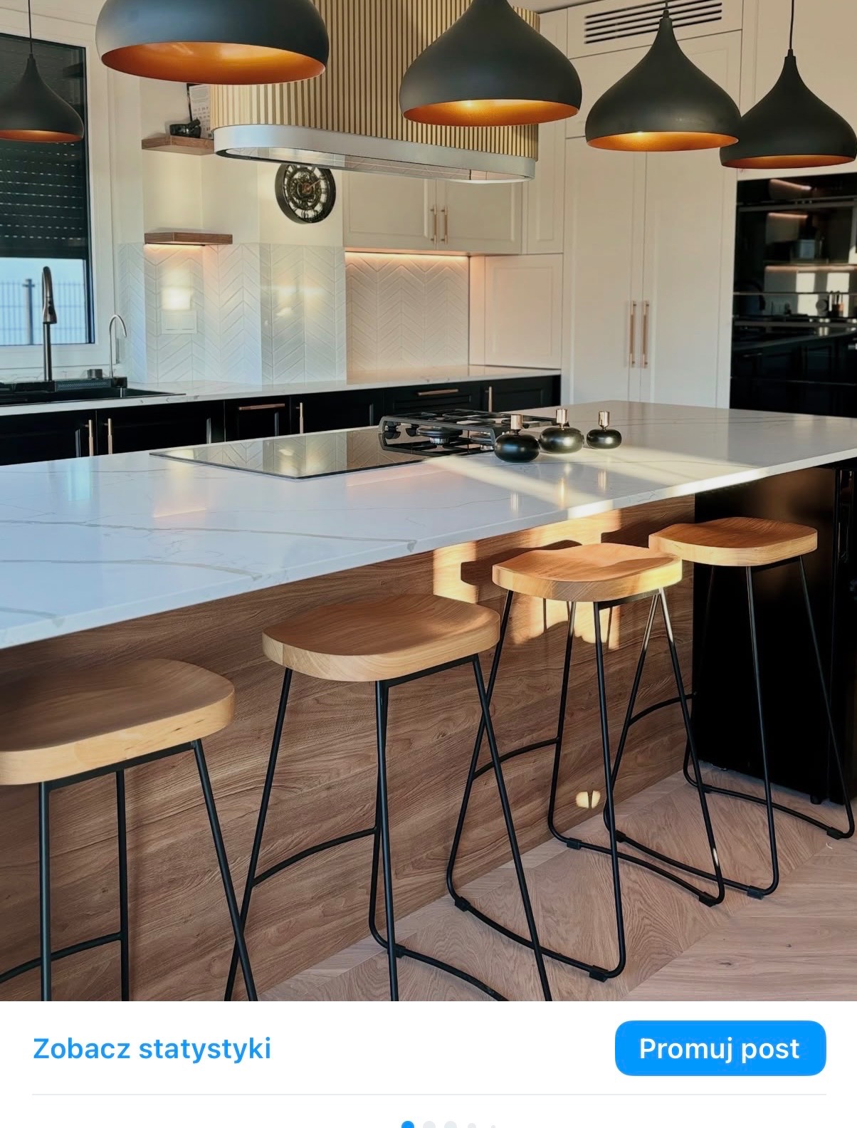 Nowoczesna kuchnia z wyspą i drewnianymi hokerami na tle białych szafek. Nad wyspą wiszą czarne lampy z miedzianym wnętrzem. Widoczny fragment okna z żaluzjami.