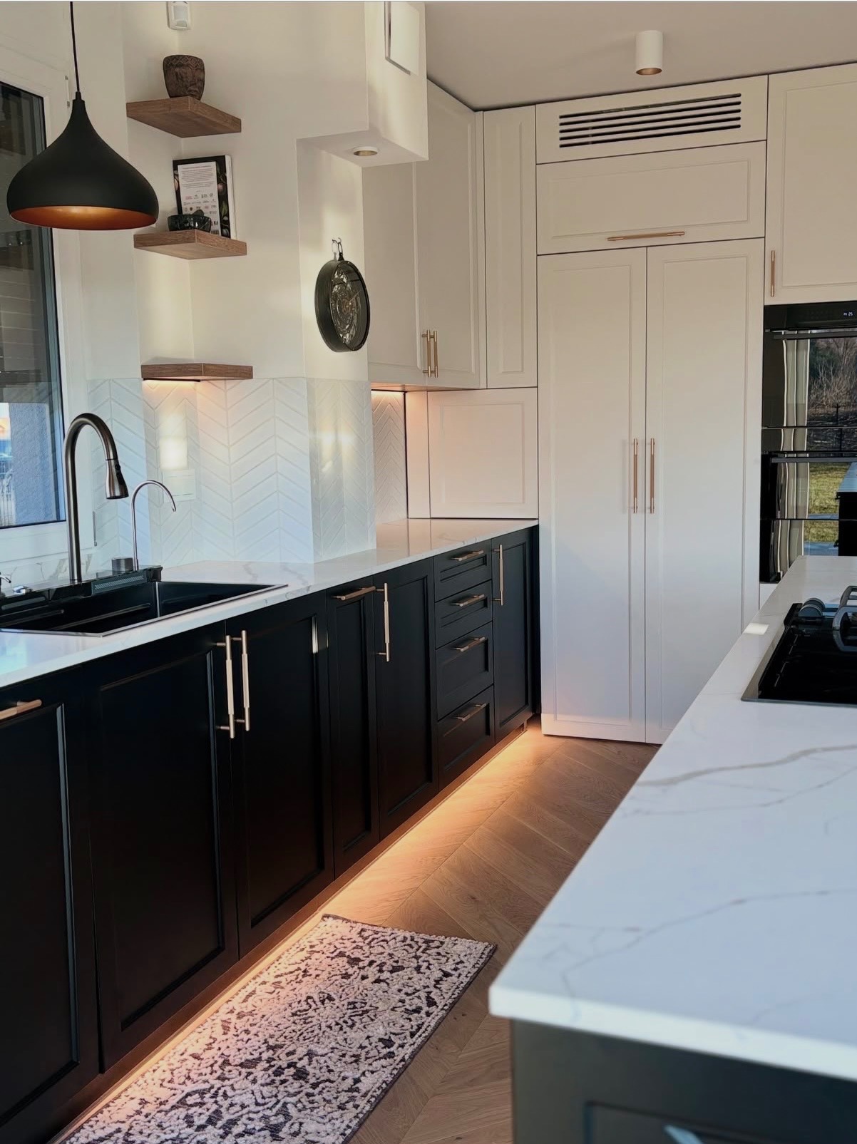 Elegancka kuchnia z czarnymi szafkami, białym blatem i złotymi uchwytami. Podświetlenie LED pod szafkami dodaje ciepła. Na ścianie dekoracyjne płytki w jodełkę.