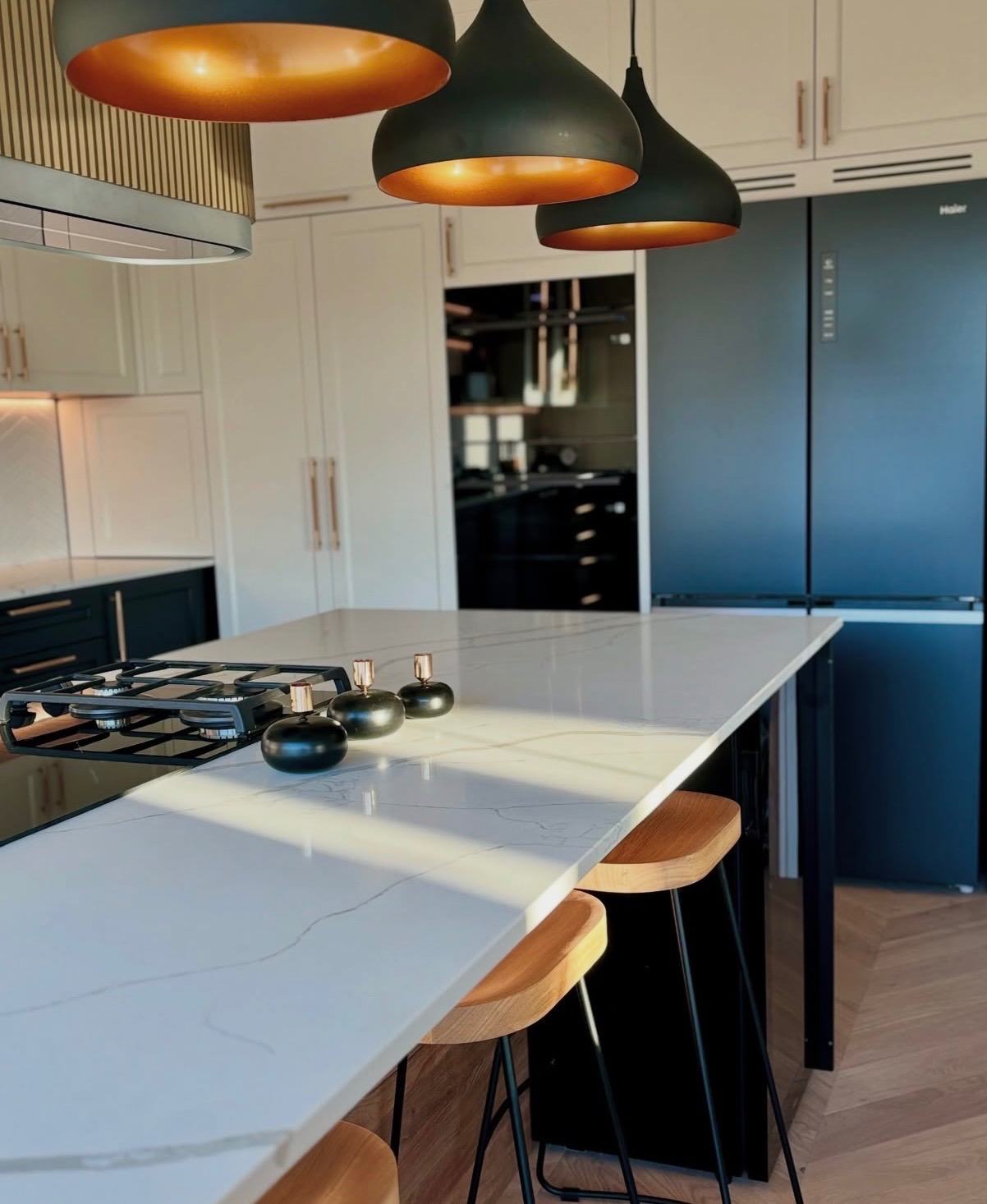 Nowoczesna kuchnia z wyspą i marmurowym blatem, oświetlona wiszącymi lampami z miedzianym wnętrzem. Widoczne drewniane hokery i czarna lodówka.