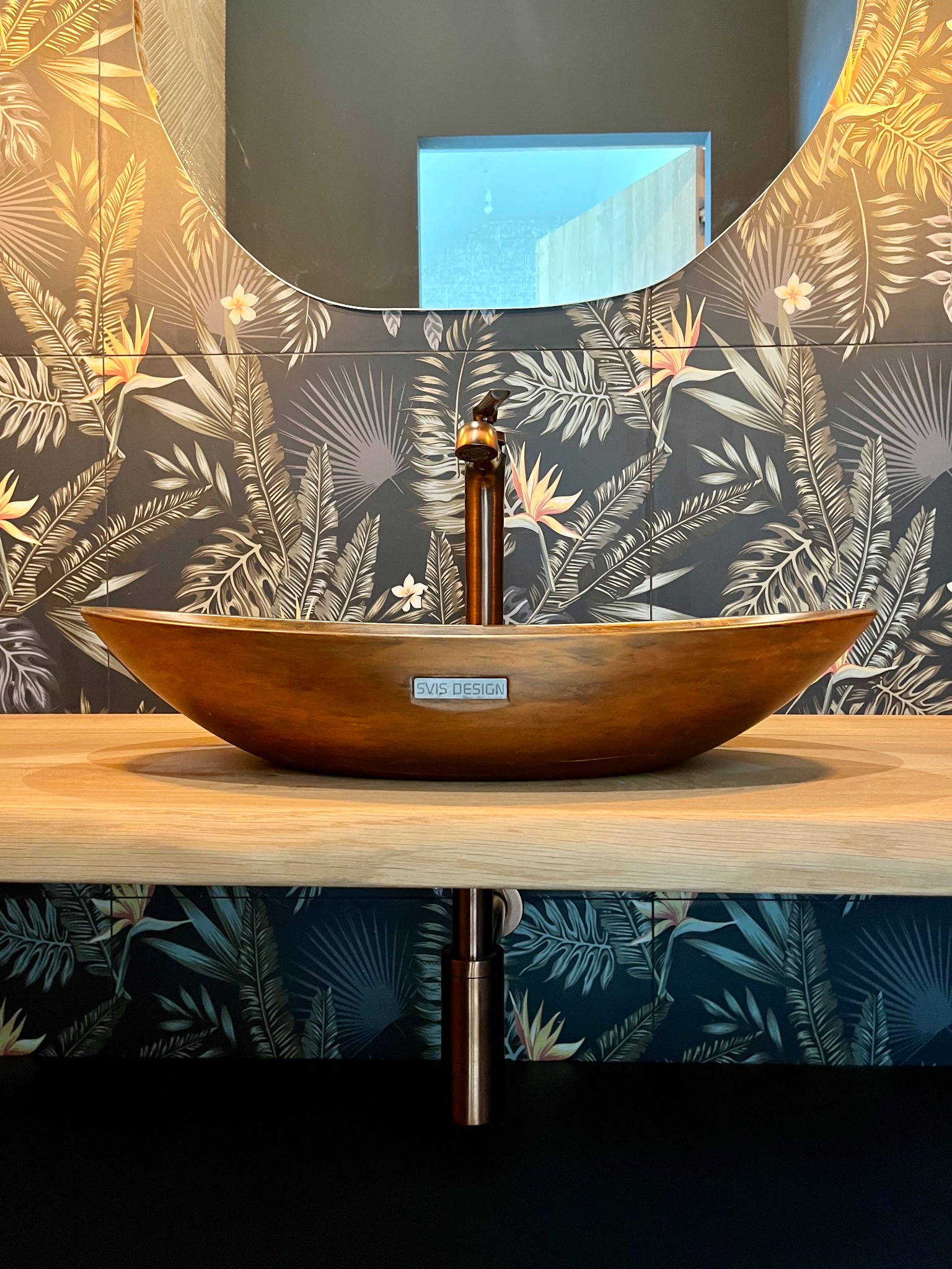Miedziana umywalka w kształcie łodzi na drewnianym blacie, z baterią w kolorze miedzi. W tle lustro i ściana w tropikalne wzory. Nowoczesna aranżacja łazienki.