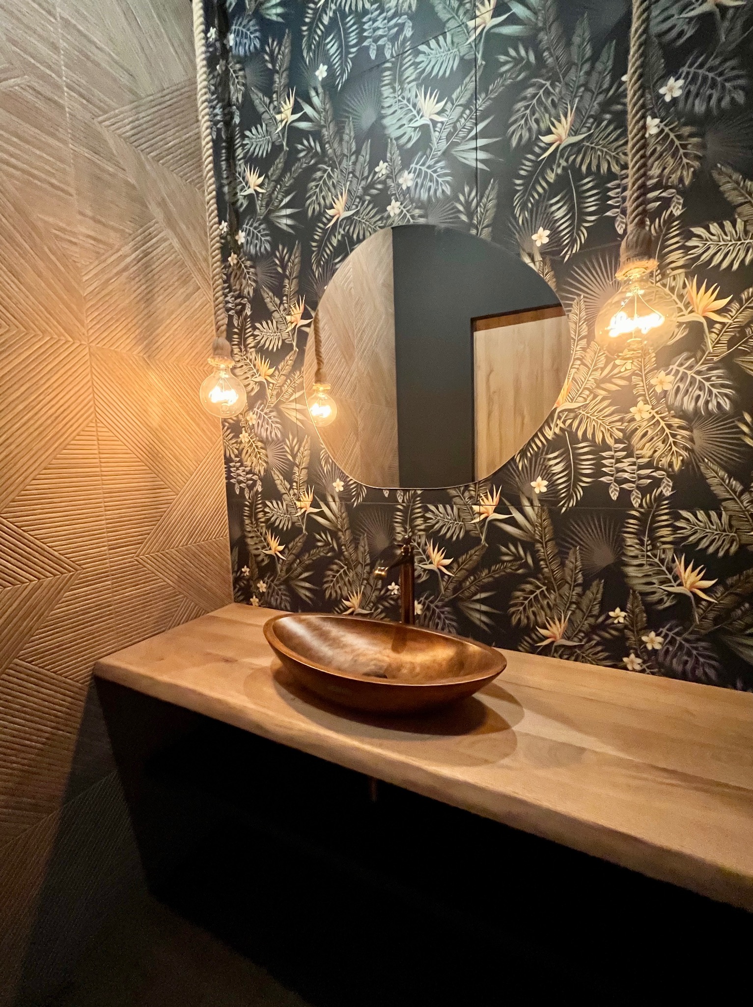 Egzotyczna łazienka: drewniany blat z miedzianą umywalką, lustro na tle ściany w tropikalne wzory, oświetlenie żarówkami na sznurach. Geometryczne płytki po lewej.