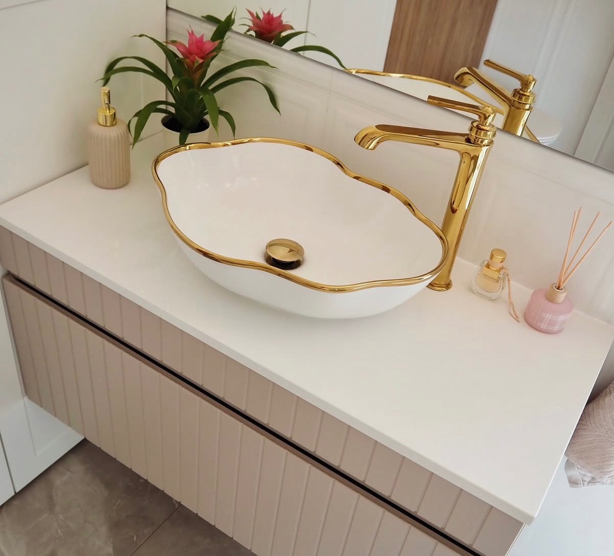 Elegancka łazienka: biała umywalka z złotym rantem i armaturą na szafce z beżowymi lamelami. W tle lustro i dekoracje, tworzące spójną, luksusową całość.