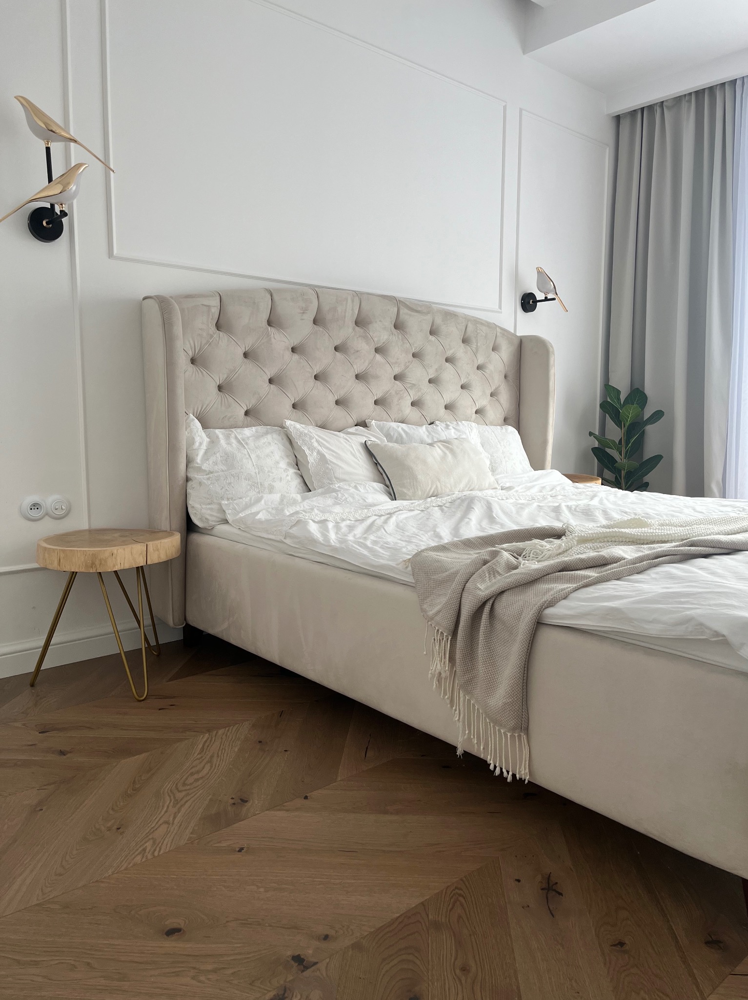 Elegancka sypialnia w bieli i beżu: tapicerowane łóżko z pikowanym zagłówkiem, drewniany stolik nocny na złotych nóżkach, parkiet w jodełkę i dekoracyjne lampki w kształcie ptaków.