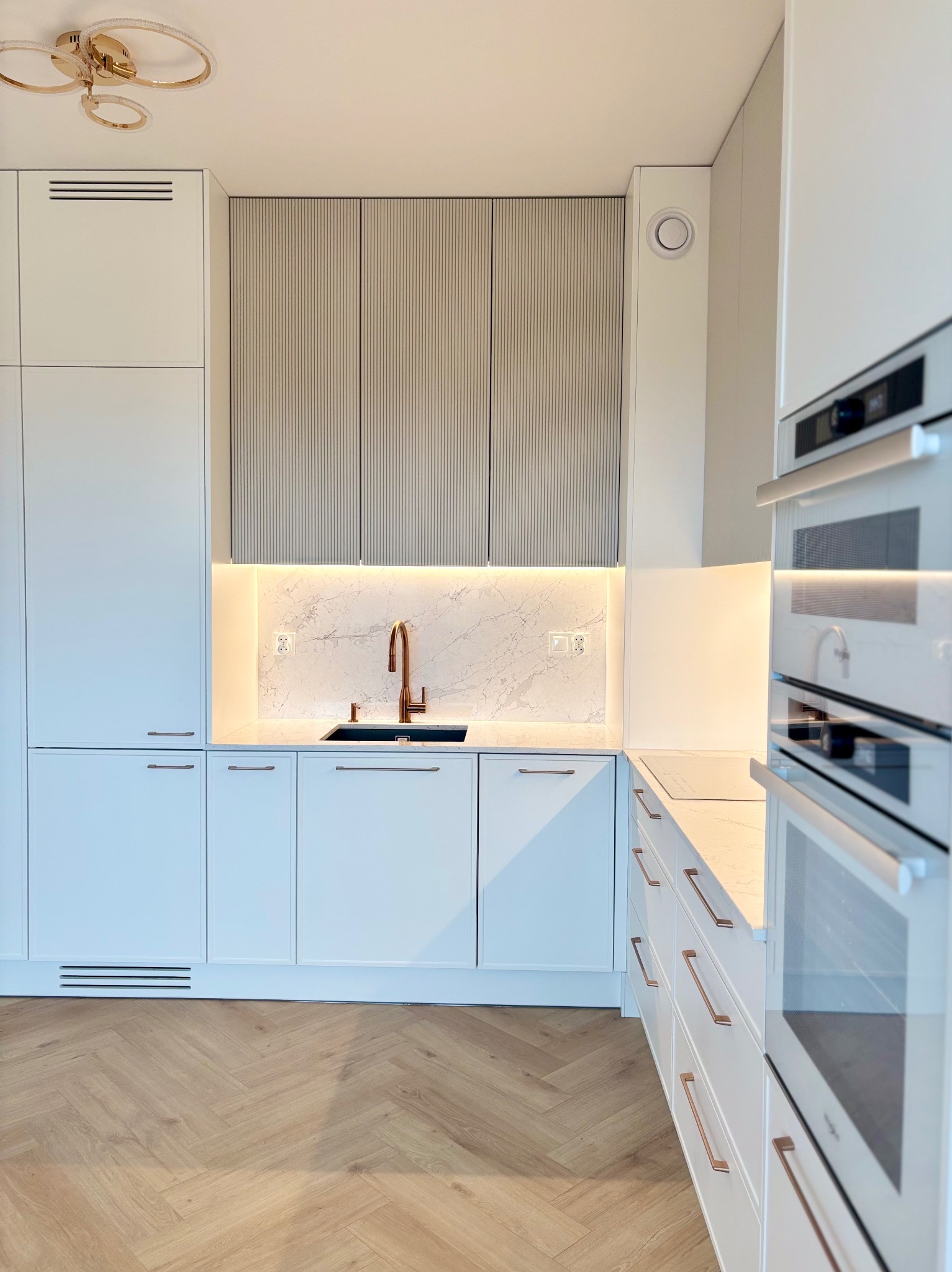 Nowoczesna kuchnia z białymi szafkami, marmurowym blatem i złotą armaturą. Parkiet w jodełkę. Oświetlenie LED podkreśla elegancję wnętrza.