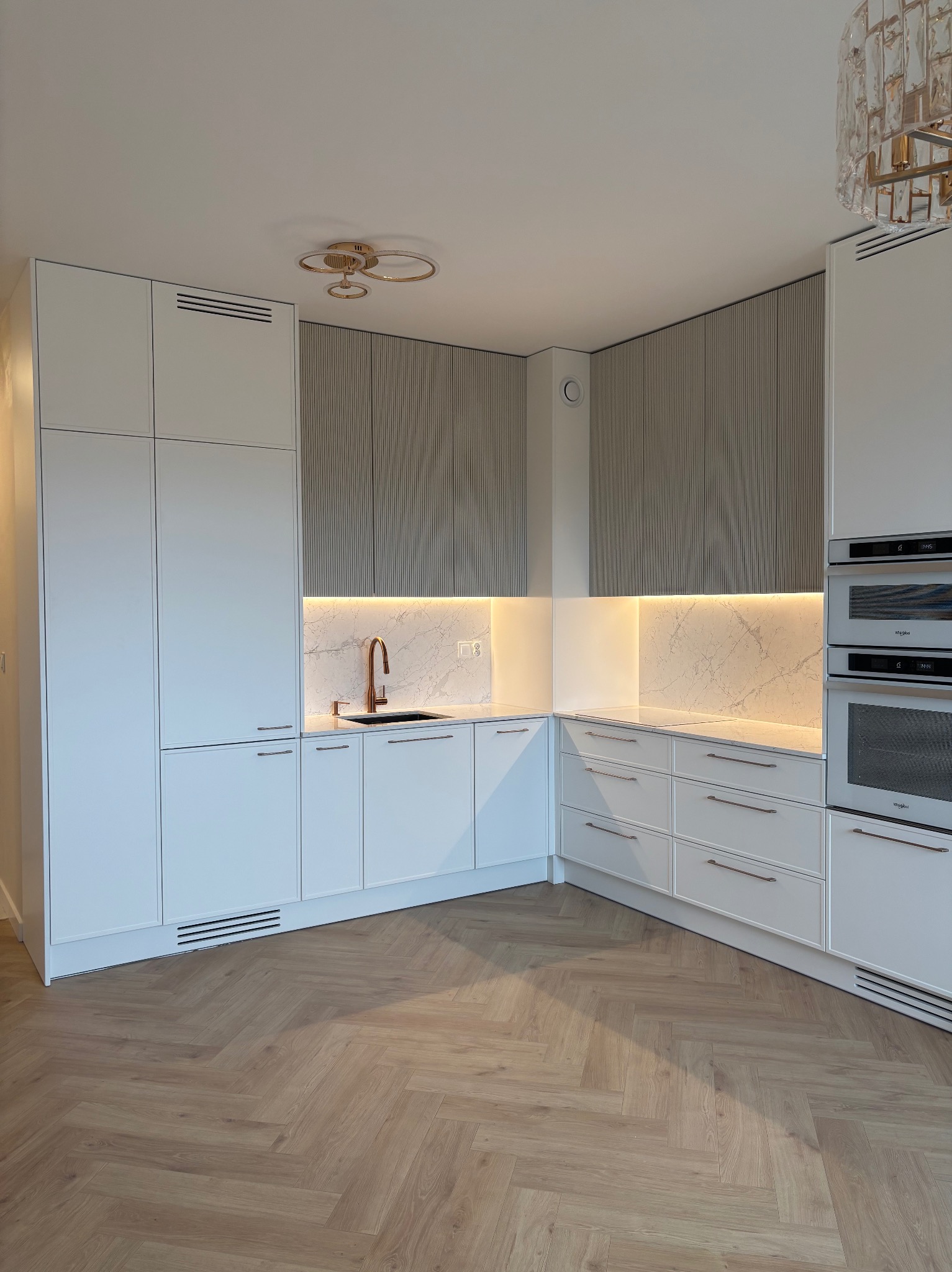 Nowoczesna kuchnia z białymi szafkami, marmurowym blatem i złotą armaturą. Parkiet w jodełkę i oświetlenie LED podkreślają elegancję wnętrza.