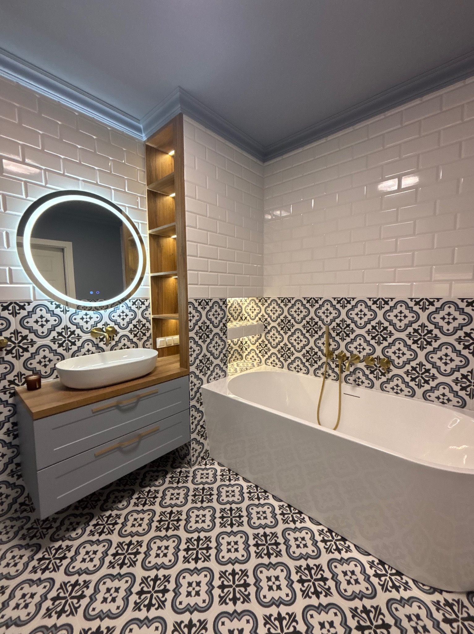 Nowoczesna łazienka z owalną umywalką na szarej szafce z drewnianym blatem, okrągłe lustro z podświetleniem LED i wolnostojąca wanna na tle wzorzystych płytek.