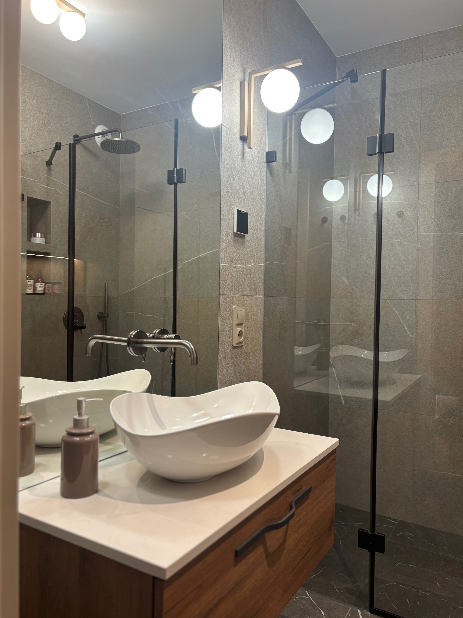Nowoczesna łazienka z szarą glazurą, kabiną prysznicową typu walk-in i umywalką nablatową. Uwagę zwraca drewniana szafka pod umywalką i designerskie oświetlenie.