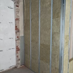 Mateusz Borowski - Ściana z płyt kartonowo-gipsowych w trakcie montażu, widoczne profile metalowe i izolacja z wełny mineralnej. Surowy stan wykończenia, fragment ściany z cegły.