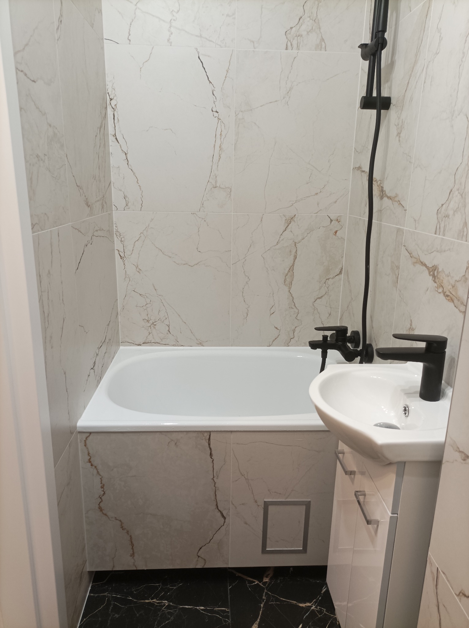 Wykończona łazienka z wanną, umywalką na szafce i czarną armaturą. Ściany i obudowa wanny wyłożone płytkami imitującymi marmur, podłoga czarna z białymi żyłkami.