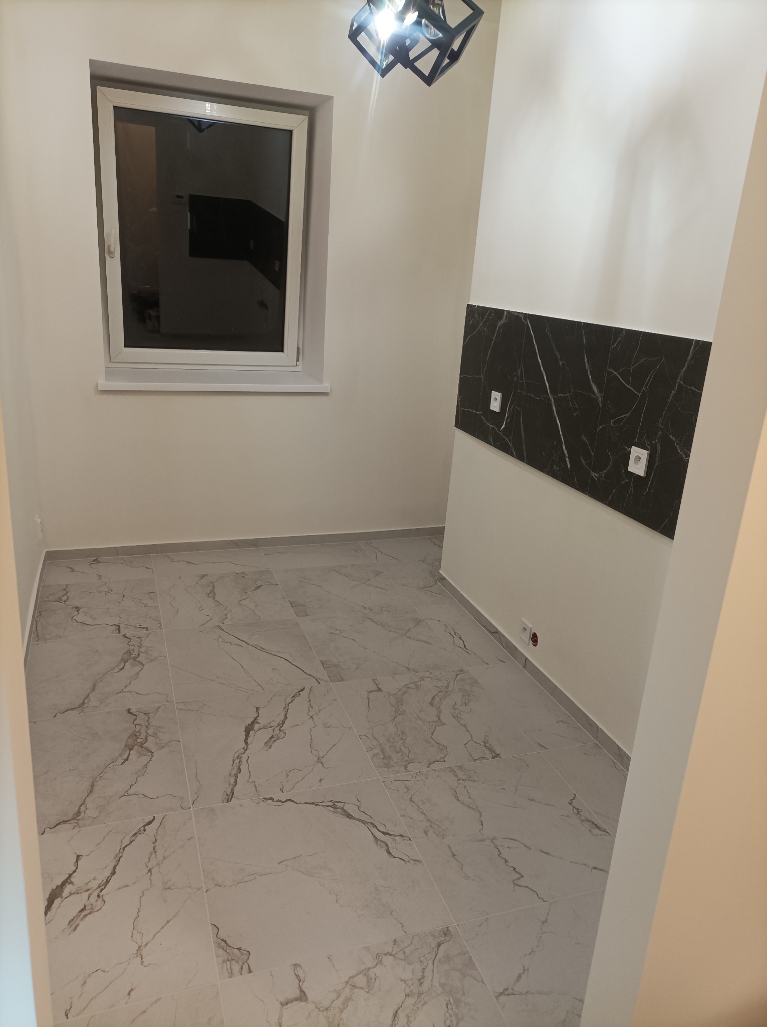 Wykończone wnętrze: marmurowe płytki podłogowe, okno z białymi ramami, dekoracyjny panel ścienny imitujący marmur z gniazdkami, nowoczesna lampa sufitowa.