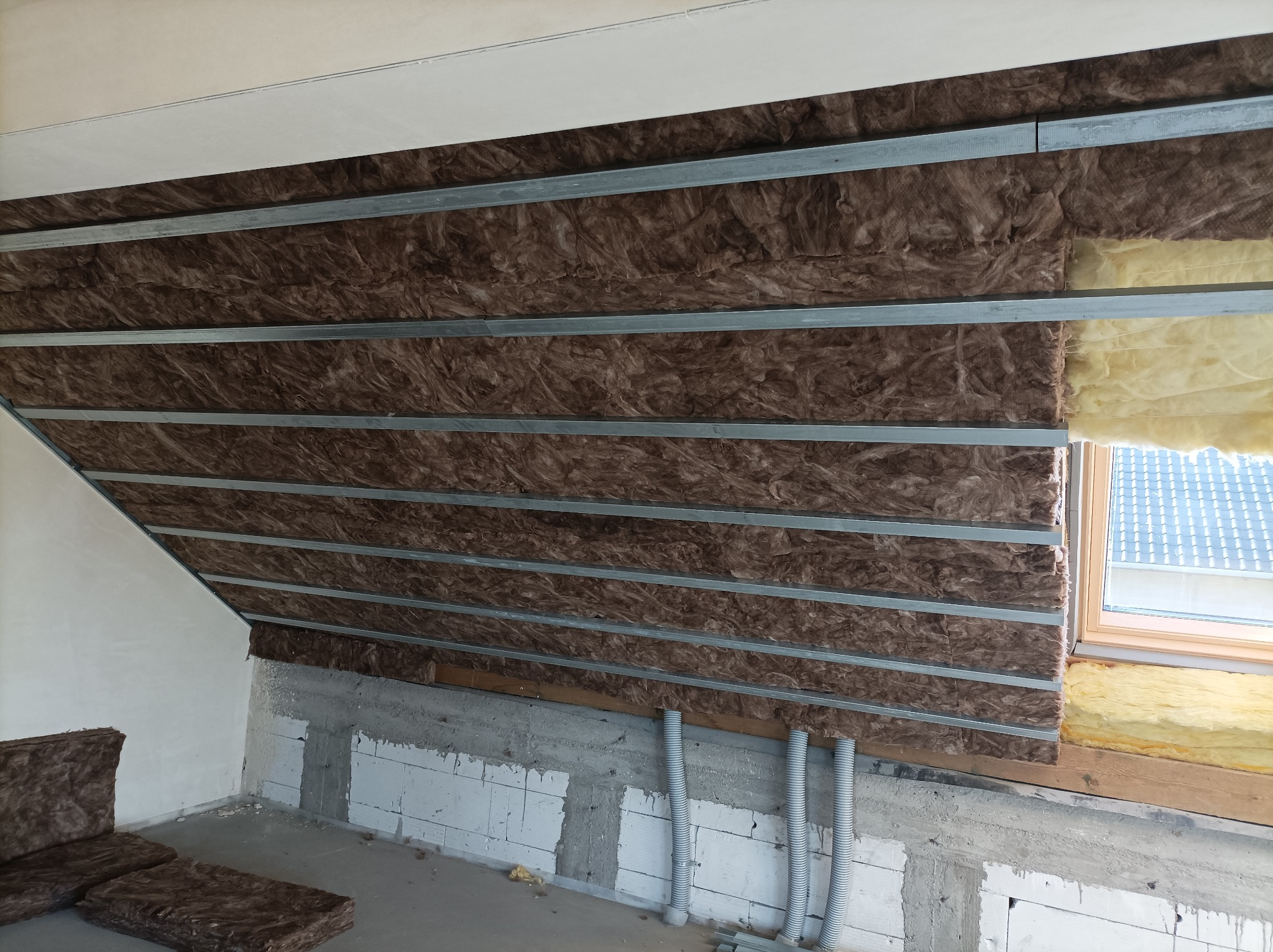 Ocieplenie poddasza wełną mineralną, widoczne metalowe profile i fragment okna dachowego. Częściowo otynkowana ściana i nieocieplona ściana z pustaków.