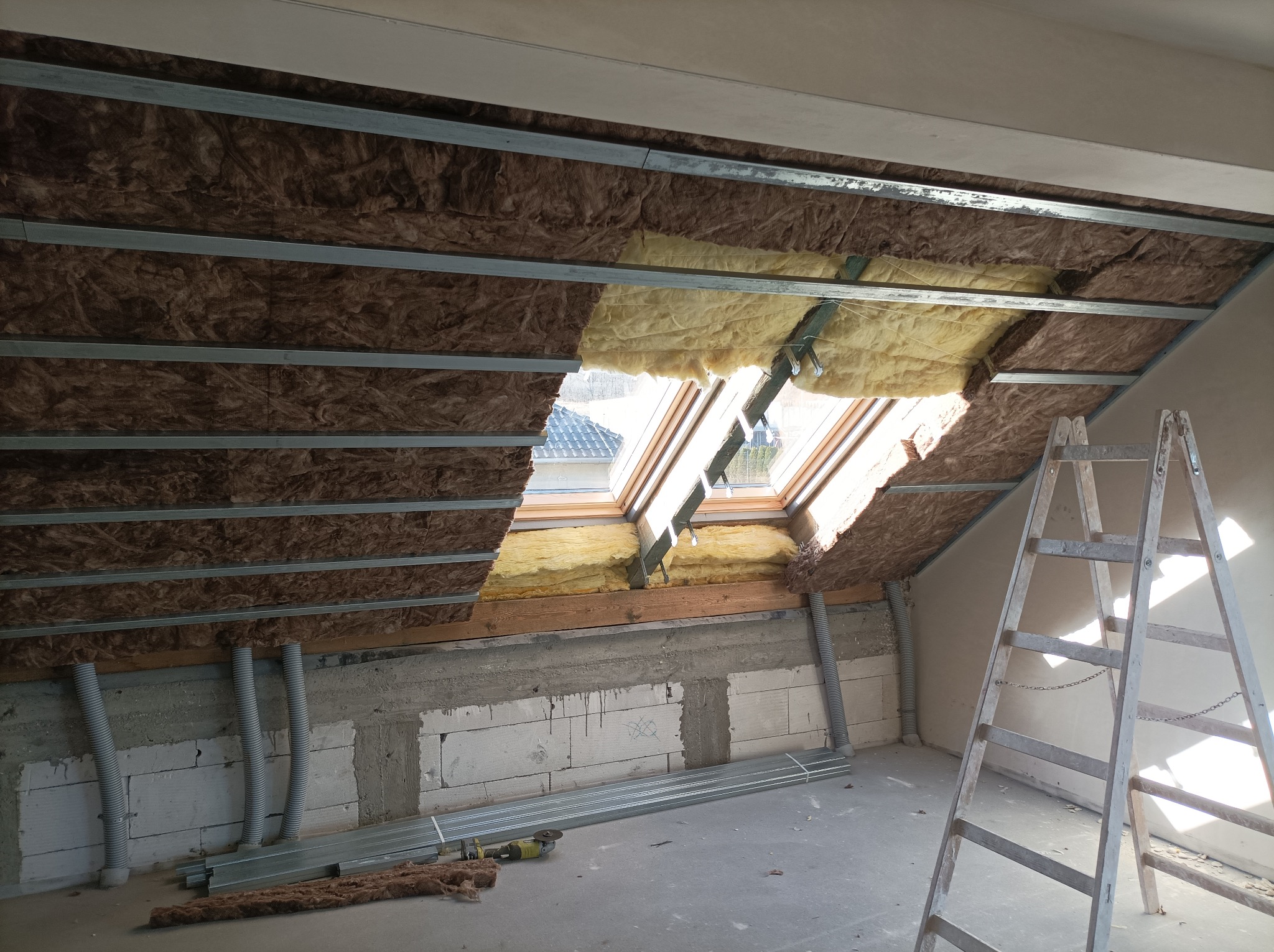 Izolacja poddasza wełną mineralną, widoczne profile metalowe i okno dachowe. Częściowo ocieplone, drabina oparta o ścianę. Prace wykończeniowe w toku.