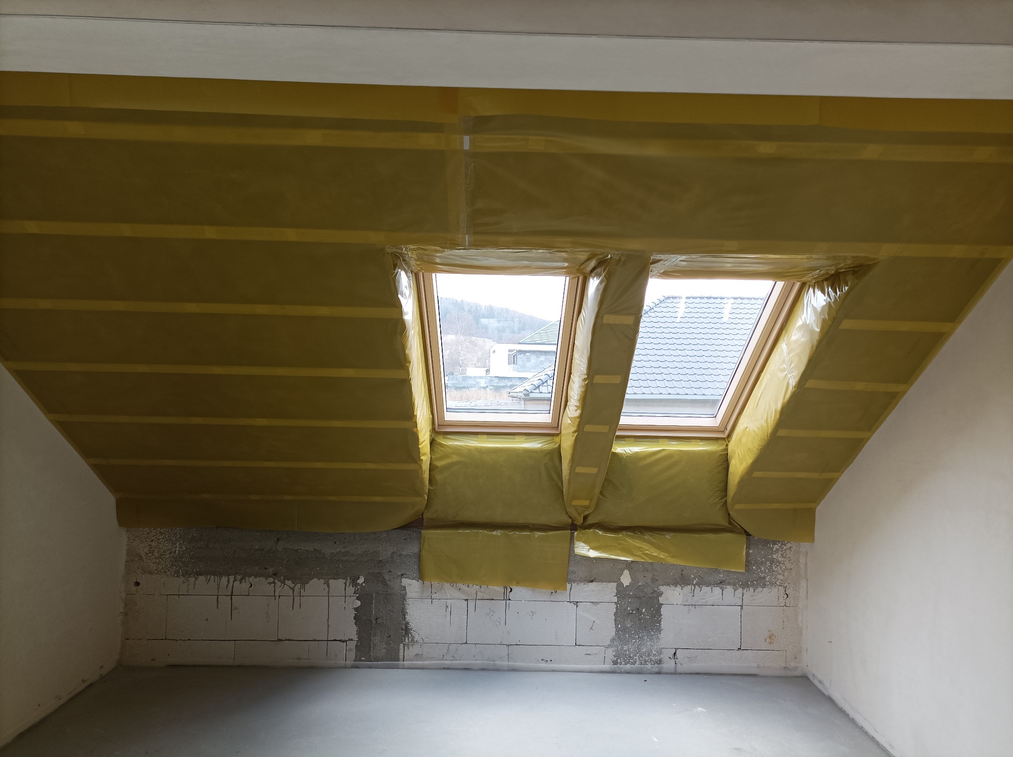 Poddasze z oknami dachowymi w trakcie izolacji. Żółta folia zabezpiecza ściany i skosy. Widoczne bloczki betonowe i surowy beton na podłodze. Etap prac wykończeniowych.