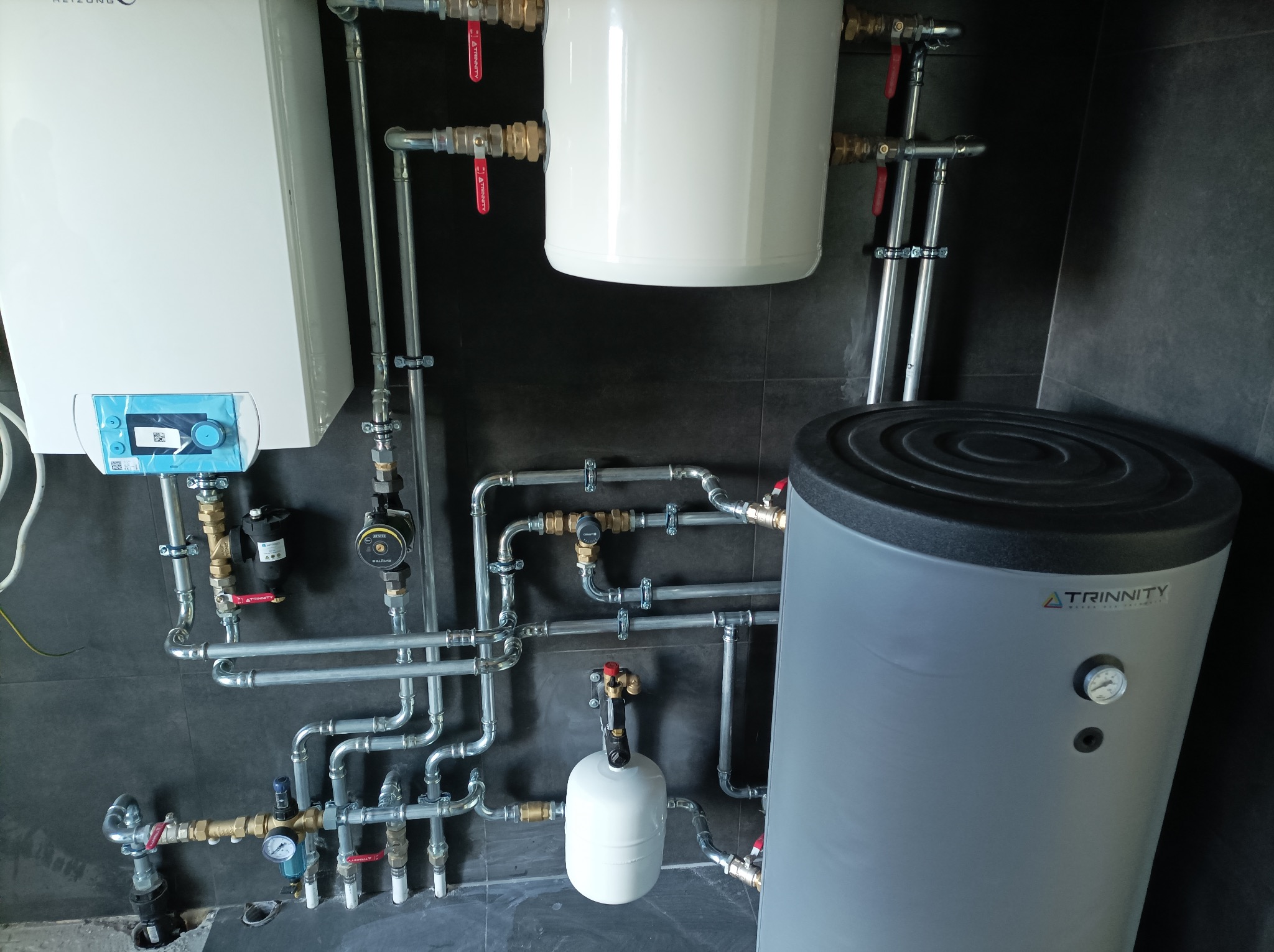 Złożona instalacja grzewcza z kotłem, zasobnikiem i rurami na tle ciemnej ściany z płytek. Widoczne zawory, pompy obiegowe i manometry. Profesjonalny montaż.