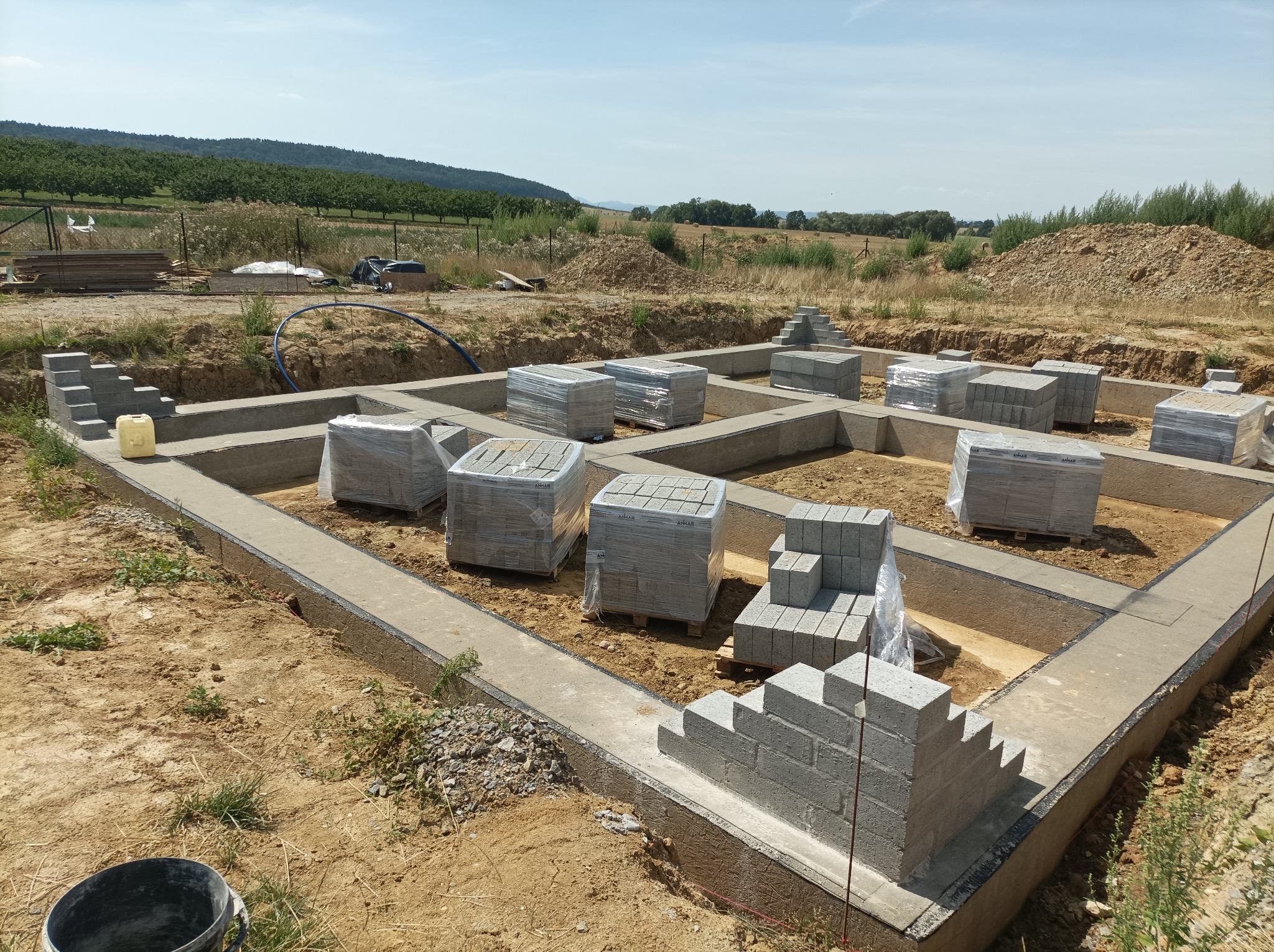 Zalane fundamenty domu jednorodzinnego z bloczków betonowych, częściowo owiniętych folią, na tle krajobrazu z drzewami i wzgórzami w słoneczny dzień.