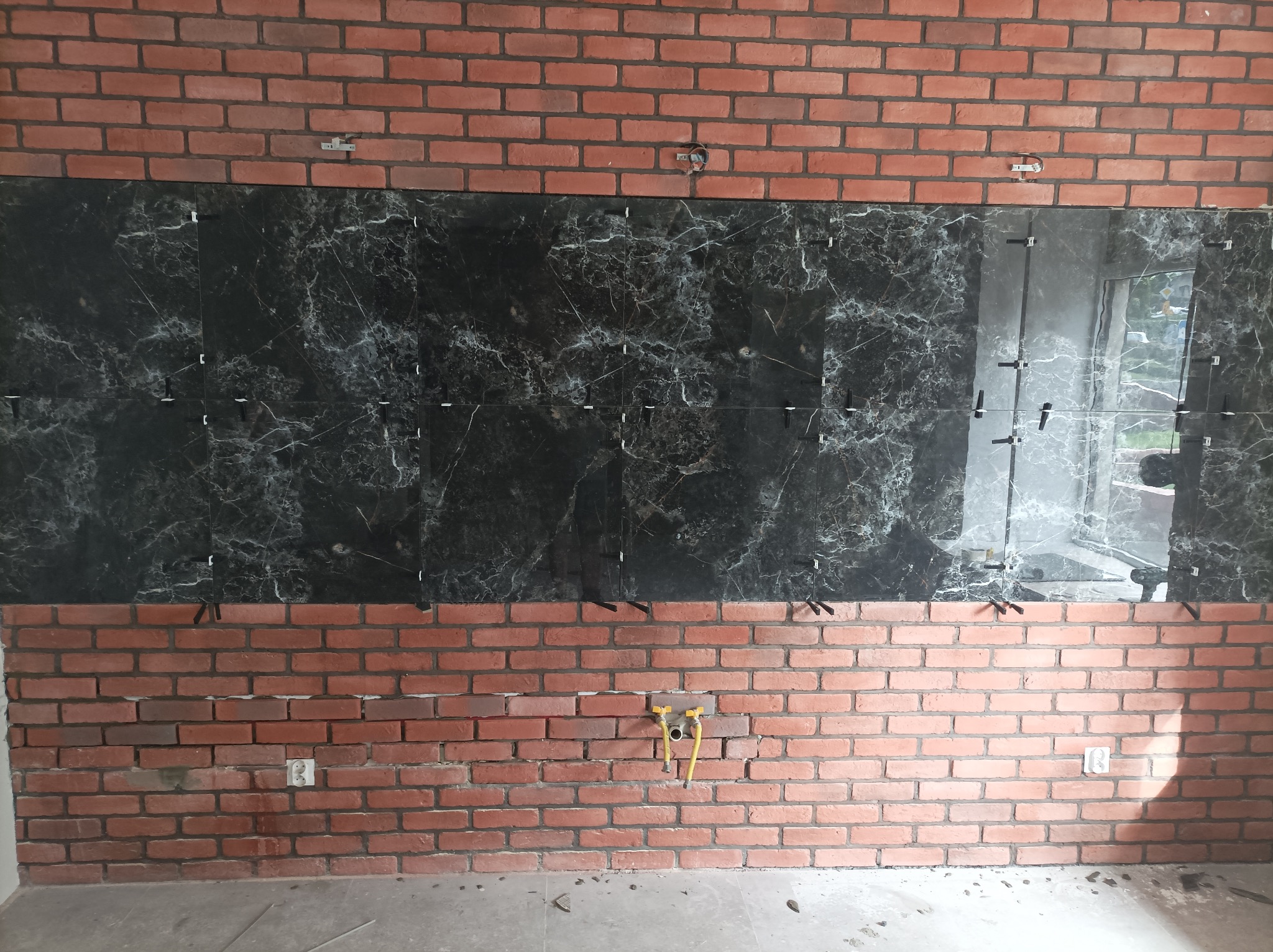 Częściowo ułożona, czarna, marmurkowa glazura na ścianie z cegły, z widocznymi krzyżykami dystansowymi i gniazdkiem elektrycznym. W tle widoczne okno.