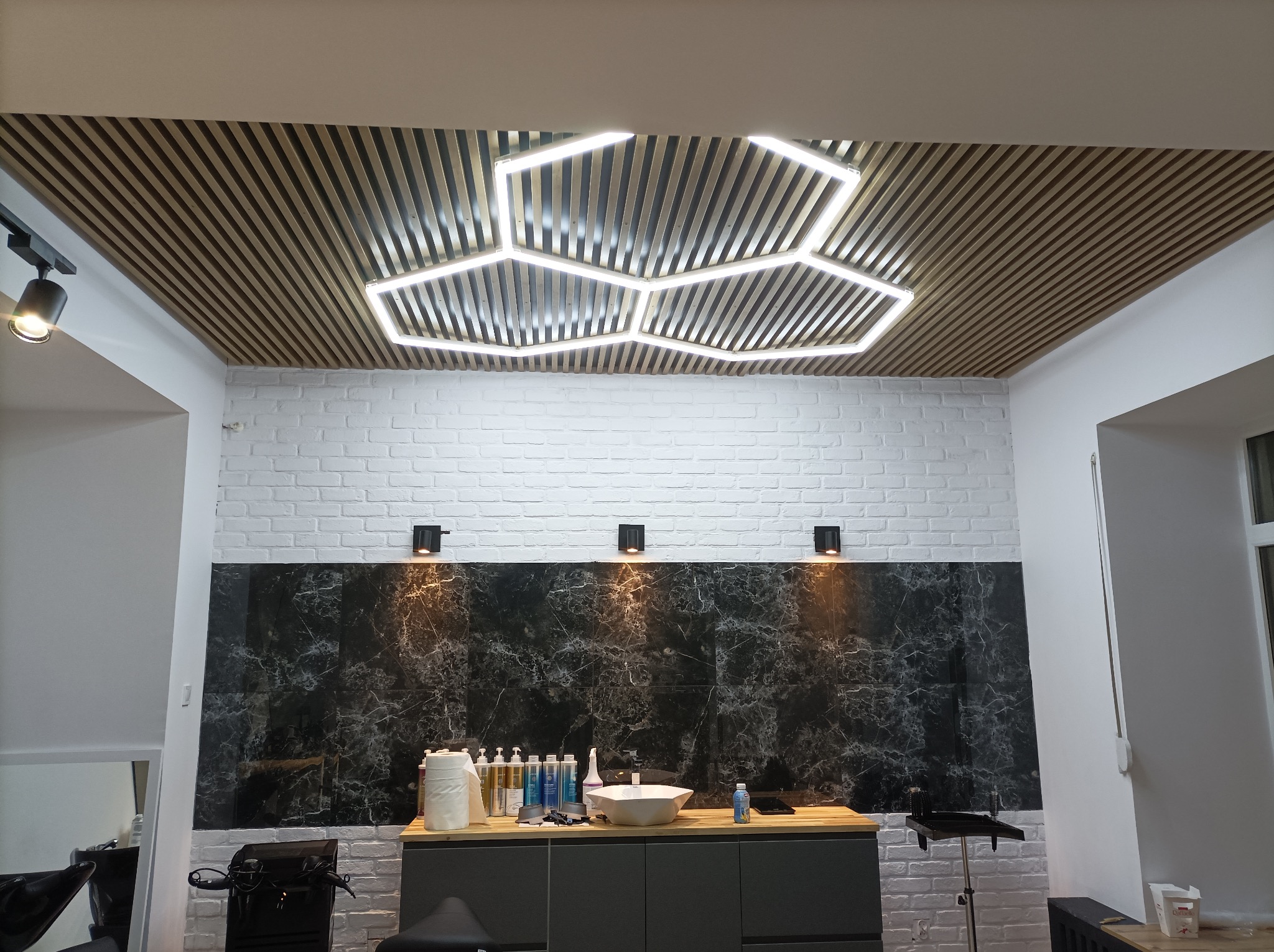 Wykończone wnętrze salonu fryzjerskiego w Wałbrzychu: drewniany sufit z heksagonalnym oświetleniem, ściana z białej cegły i marmurowy panel nad umywalką.