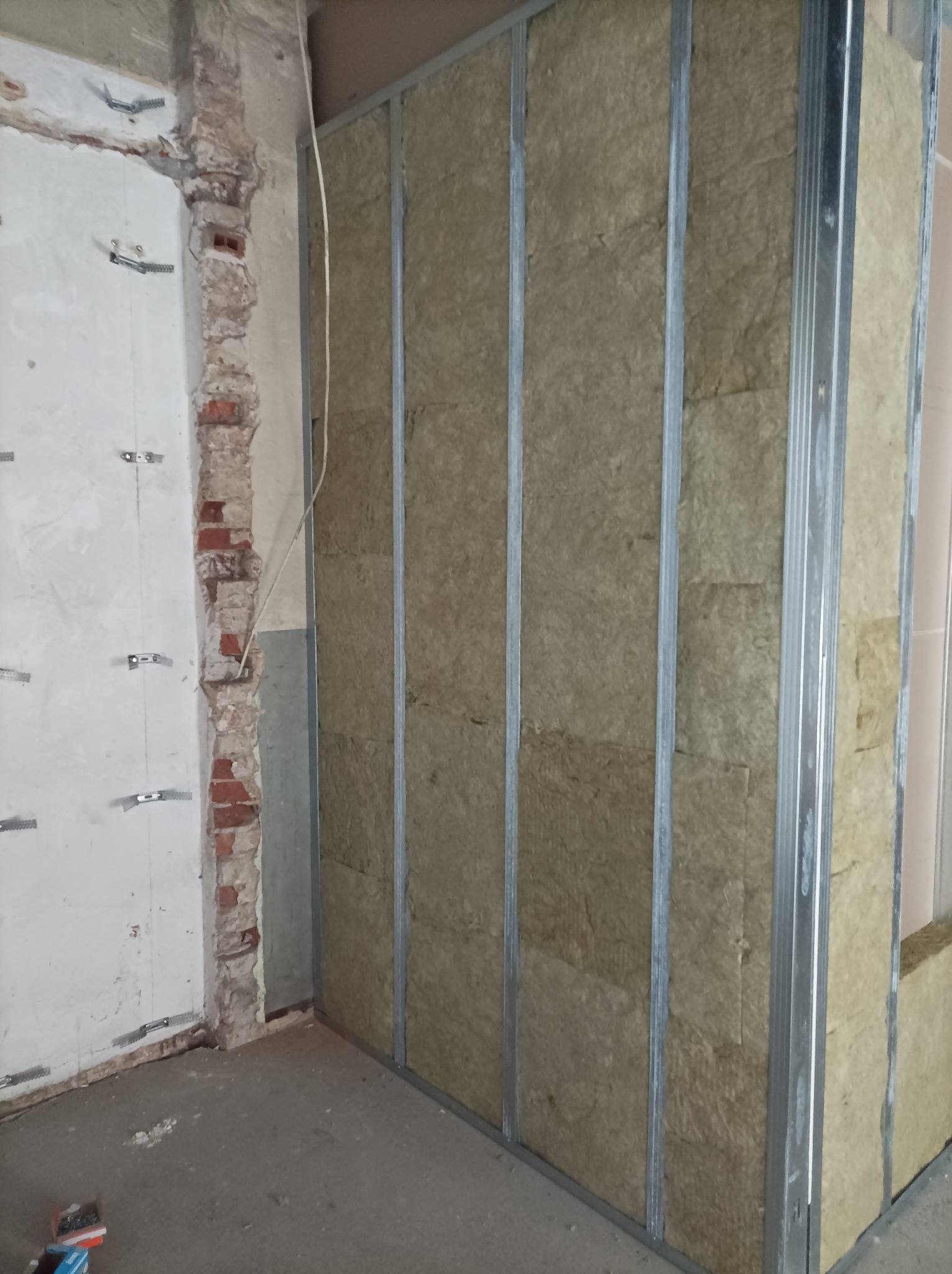 Ściana z płyt kartonowo-gipsowych w trakcie montażu, widoczne profile metalowe i izolacja z wełny mineralnej. Surowy stan wykończenia, fragment ściany z cegły.