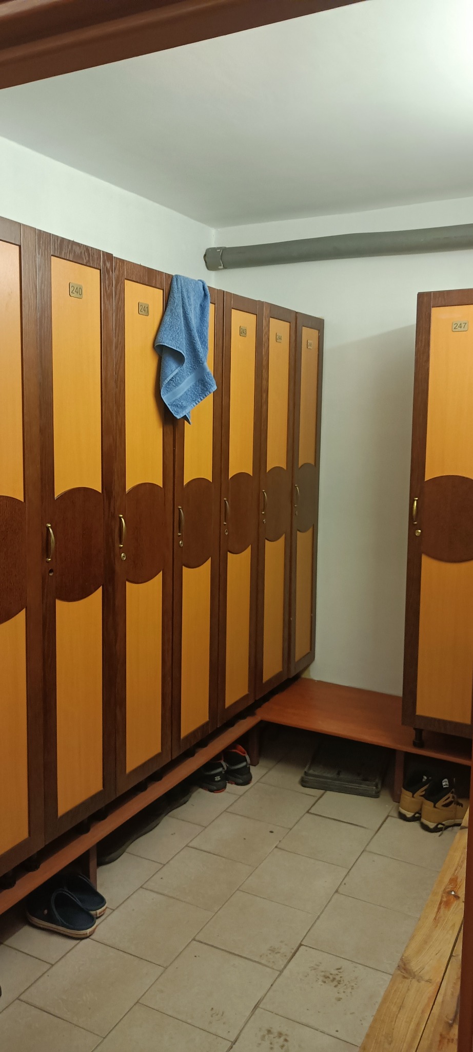 Szereg drewnianych szafek z numerami w szatni, z niebieskim ręcznikiem wiszącym na jednej z nich. Pod szafkami widoczne buty.