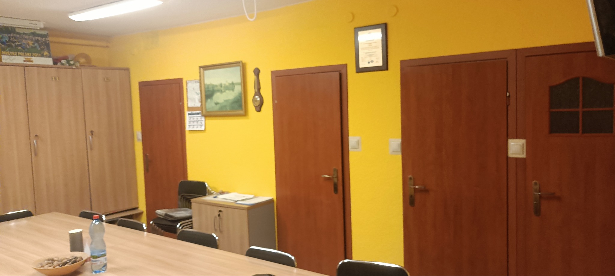 Żółta ściana po odświeżeniu w biurze w Gdyni. Widoczne drzwi, obraz, szafki i stół konferencyjny. Ściana pomalowana na jednolity, intensywny kolor.