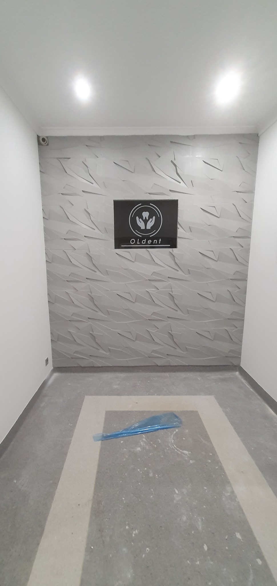 Wnętrze z dekoracyjną ścianą 3D i logo 'Oldent' w czarnej ramce. Podłoga w szaro-białe wzory, zabezpieczona folią. Minimalistyczny design, oświetlenie punktowe.