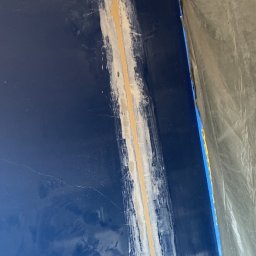 Aqua Rem Firma Remontowo-Budowlana - Bruzdy pod kable elektryczne wypełnione gipsem na niebieskiej ścianie, widoczne otwory pod gniazdka, zabezpieczona folią część pomieszczenia.