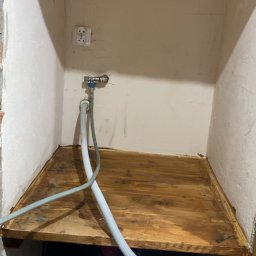 Aqua Rem Firma Remontowo-Budowlana - Niewielka wnęka z drewnianą półką, widoczny zawór wodny, gniazdko elektryczne i fragment pralki. Ujęcie z dołu, podkreślające ciasnotę przestrzeni.