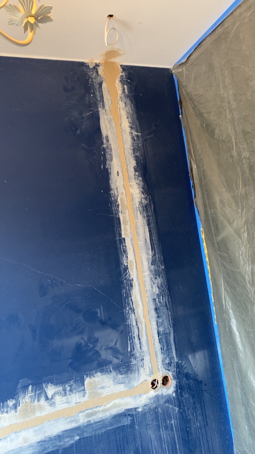 Bruzdy pod kable elektryczne wypełnione gipsem na niebieskiej ścianie, widoczne otwory pod gniazdka, zabezpieczona folią część pomieszczenia.