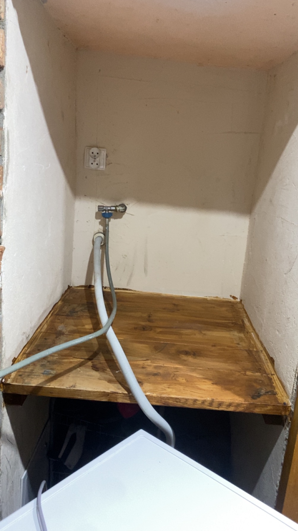 Niewielka wnęka z drewnianą półką, widoczny zawór wodny, gniazdko elektryczne i fragment pralki. Ujęcie z dołu, podkreślające ciasnotę przestrzeni.