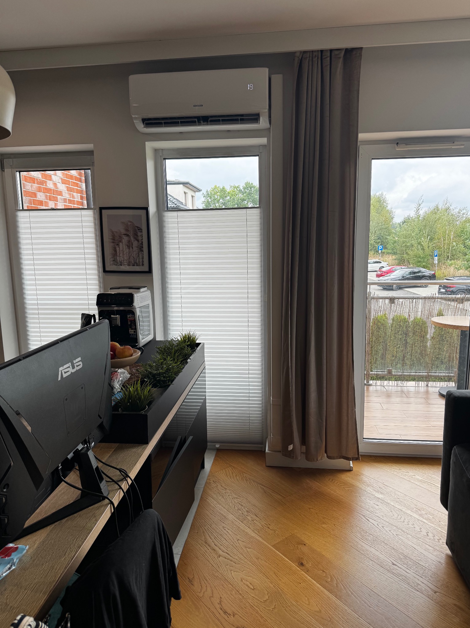 Biały klimatyzator ścienny zamontowany nad oknem z żaluzjami w jasnym pokoju z wyjściem na balkon, widoczne zasłony i elementy wyposażenia wnętrza.