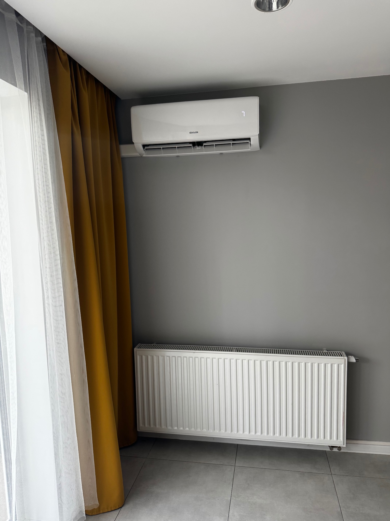 Biały klimatyzator ścienny nad grzejnikiem w szarym pokoju z zasłonami. Widok na zamontowane urządzenie w nowoczesnym wnętrzu.