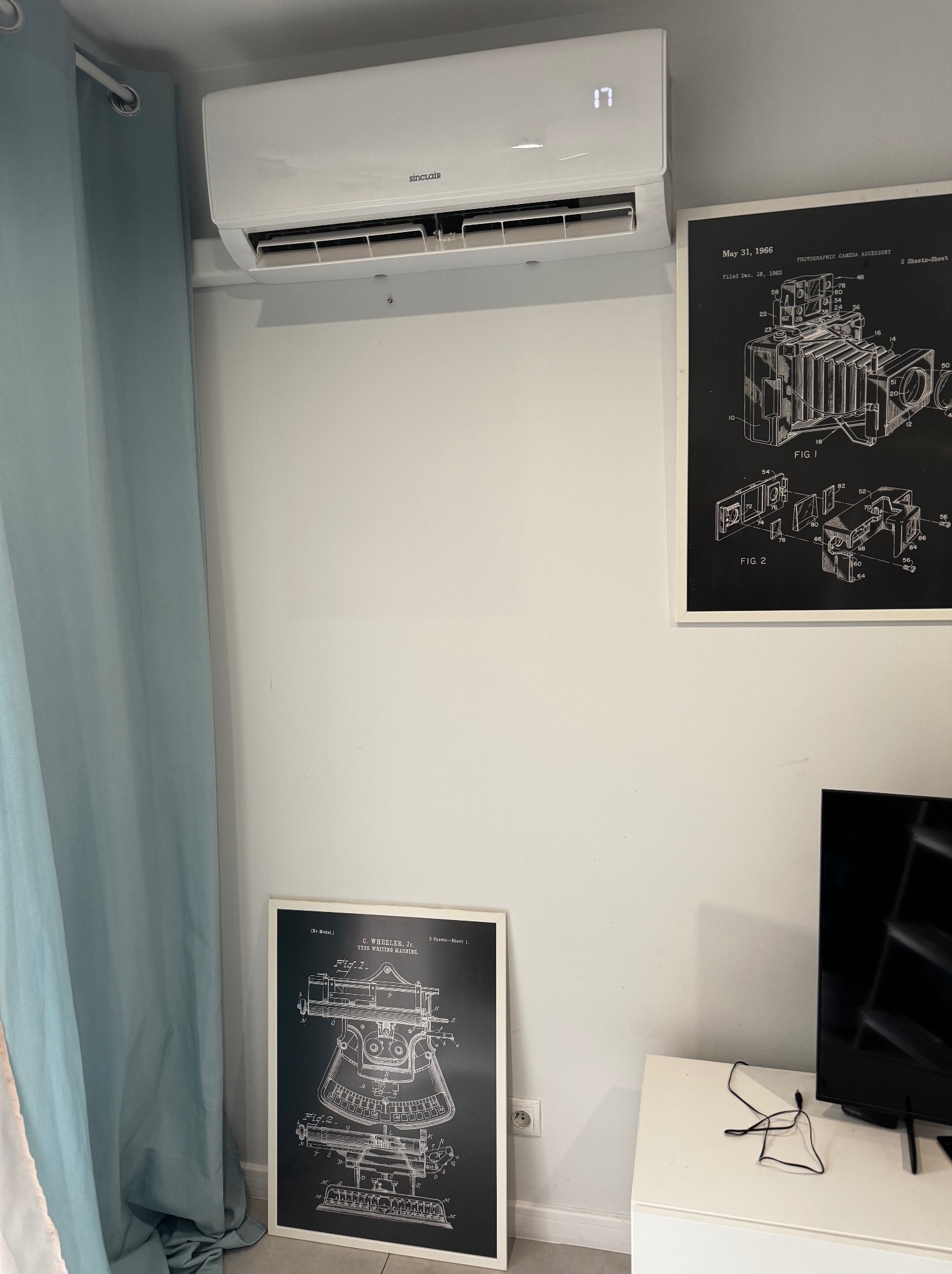 Biały klimatyzator Sinclair na ścianie w pokoju, obok wiszą grafiki w ramkach. Widoczny fragment zasłony i telewizora. Nowoczesny design, minimalistyczne wnętrze.