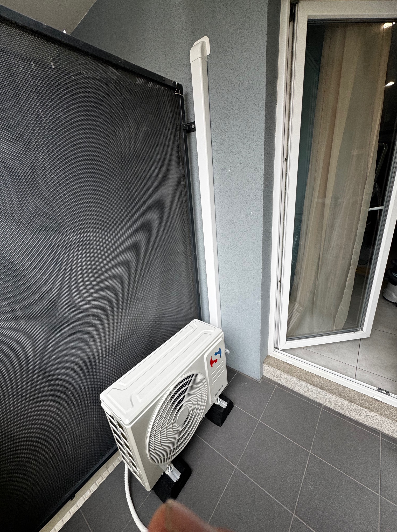 Biała jednostka zewnętrzna klimatyzacji zamontowana na szarym balkonie, widoczna czarna osłona i otwarte drzwi balkonowe. Minimalistyczny design.