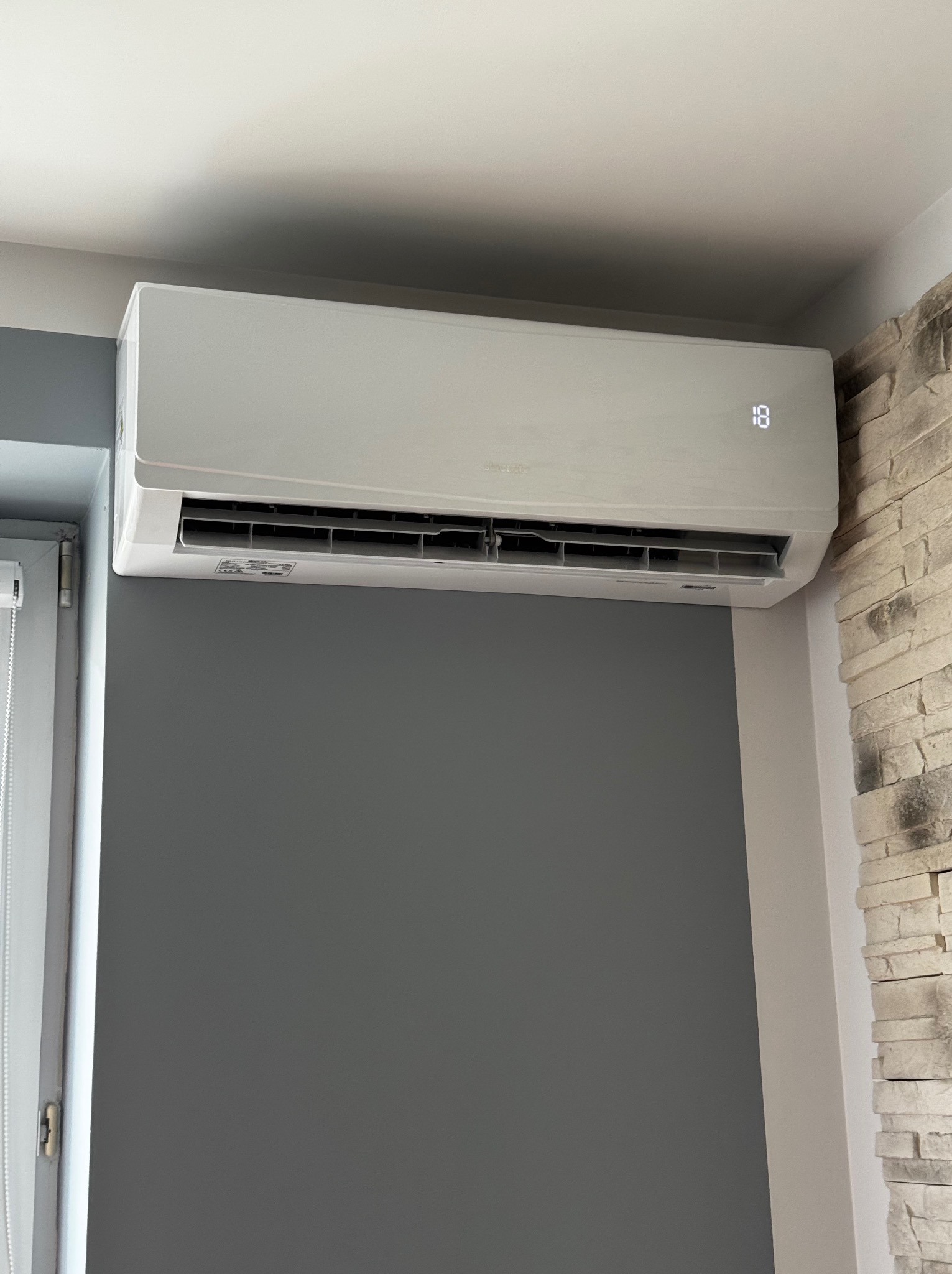 Biały klimatyzator ścienny z wyświetlaczem temperatury (18°C), zamontowany w rogu pokoju z szarą ścianą i dekoracyjną ścianą z kamienia.