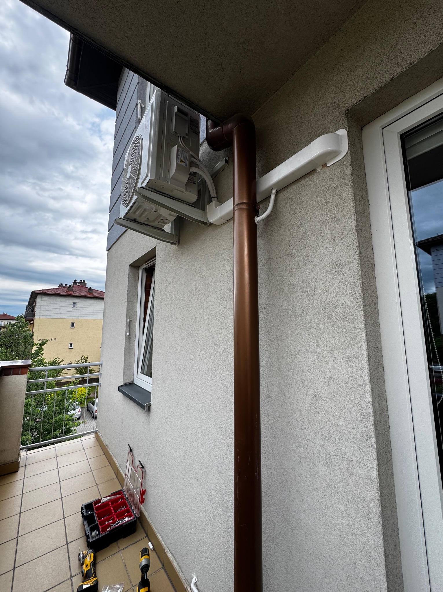 Zamontowany klimatyzator na elewacji budynku, widoczna rura odprowadzająca skropliny. Na balkonie narzędzia montera. Ujęcie z dołu, pochmurne niebo w tle.