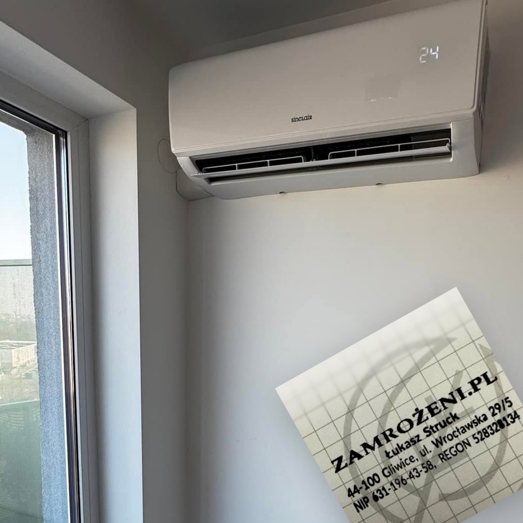 Biały klimatyzator Sinclair zamontowany na ścianie, obok okna. W rogu wizytówka firmy z adresem i numerem NIP. Widoczny wyświetlacz z temperaturą 24 stopnie.
