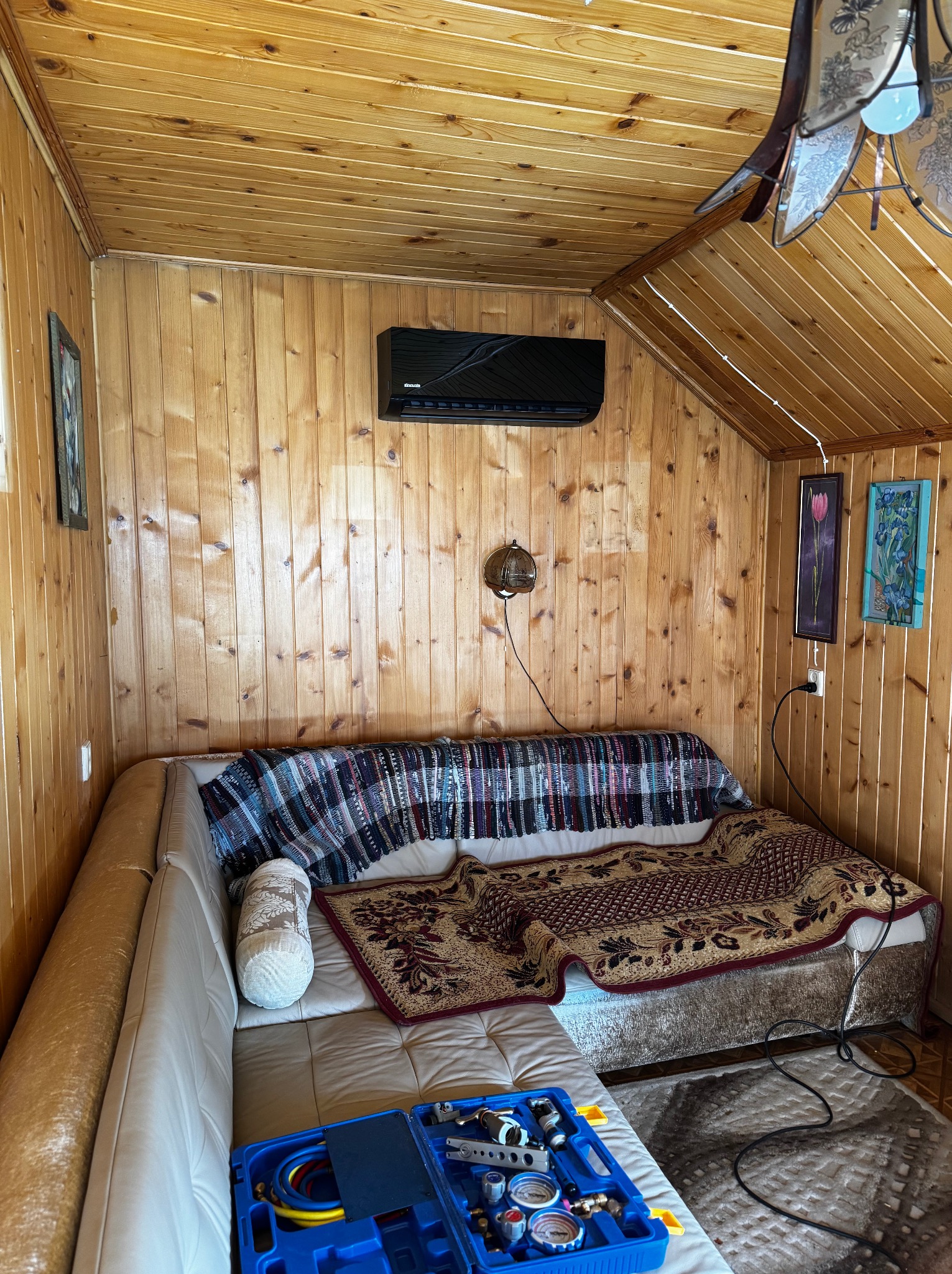 Klimatyzator ścienny w drewnianym wnętrzu, nad sofą. Na pierwszym planie narzędzia w niebieskiej skrzynce. Widoczne przewody i manometry.