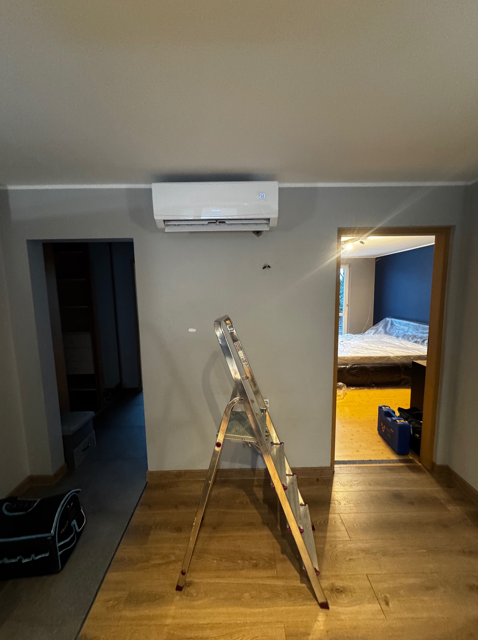 Biały klimatyzator na szarej ścianie w pokoju, widoczna drabina i torba narzędziowa, sugerujące świeży montaż. W tle widok na sypialnię.