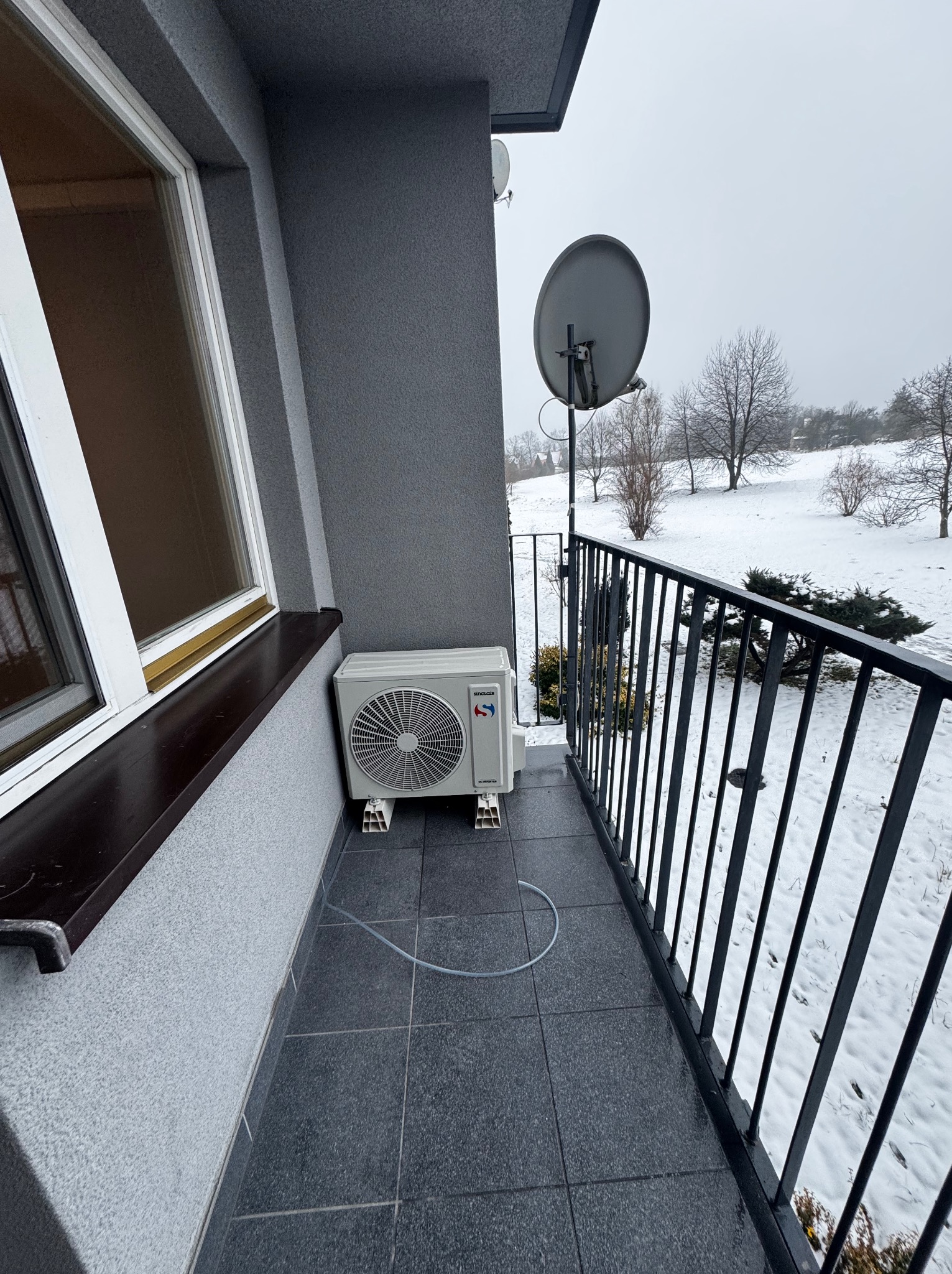 Klimatyzator na balkonie z widokiem na zimowy krajobraz. Jednostka zewnętrzna klimatyzacji zamontowana przy ścianie budynku. Widoczna antena satelitarna w tle.