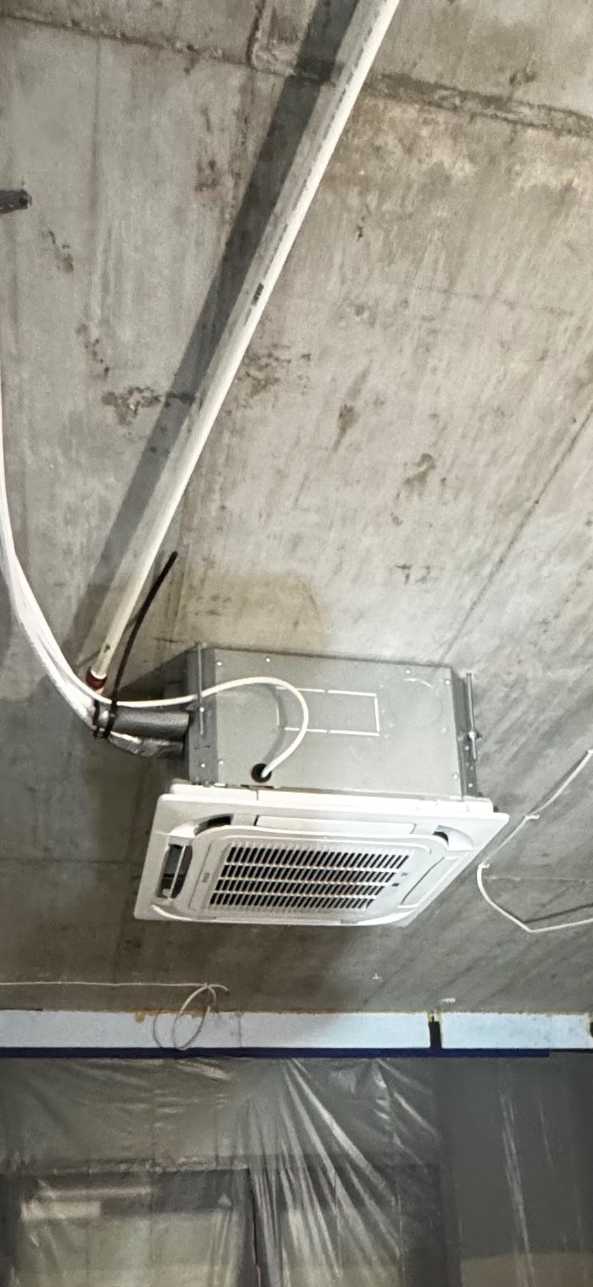 Kasetonowa jednostka klimatyzacji zamontowana pod betonowym sufitem, widoczne rury instalacyjne i zabezpieczenie folią malarską. Ujęcie z dołu.