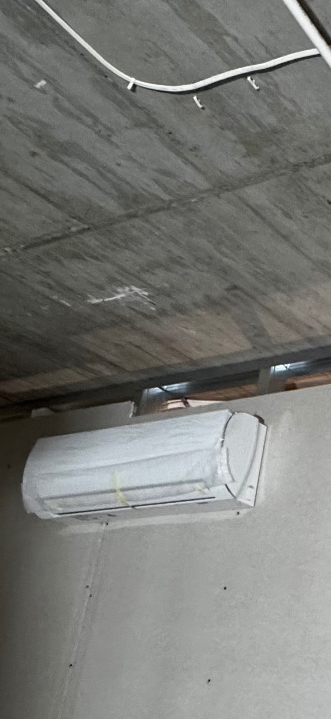 Zamontowany klimatyzator na ścianie w surowym wnętrzu. Widoczne przewody pod sufitem i niedokończone ściany. Urządzenie zabezpieczone folią ochronną.