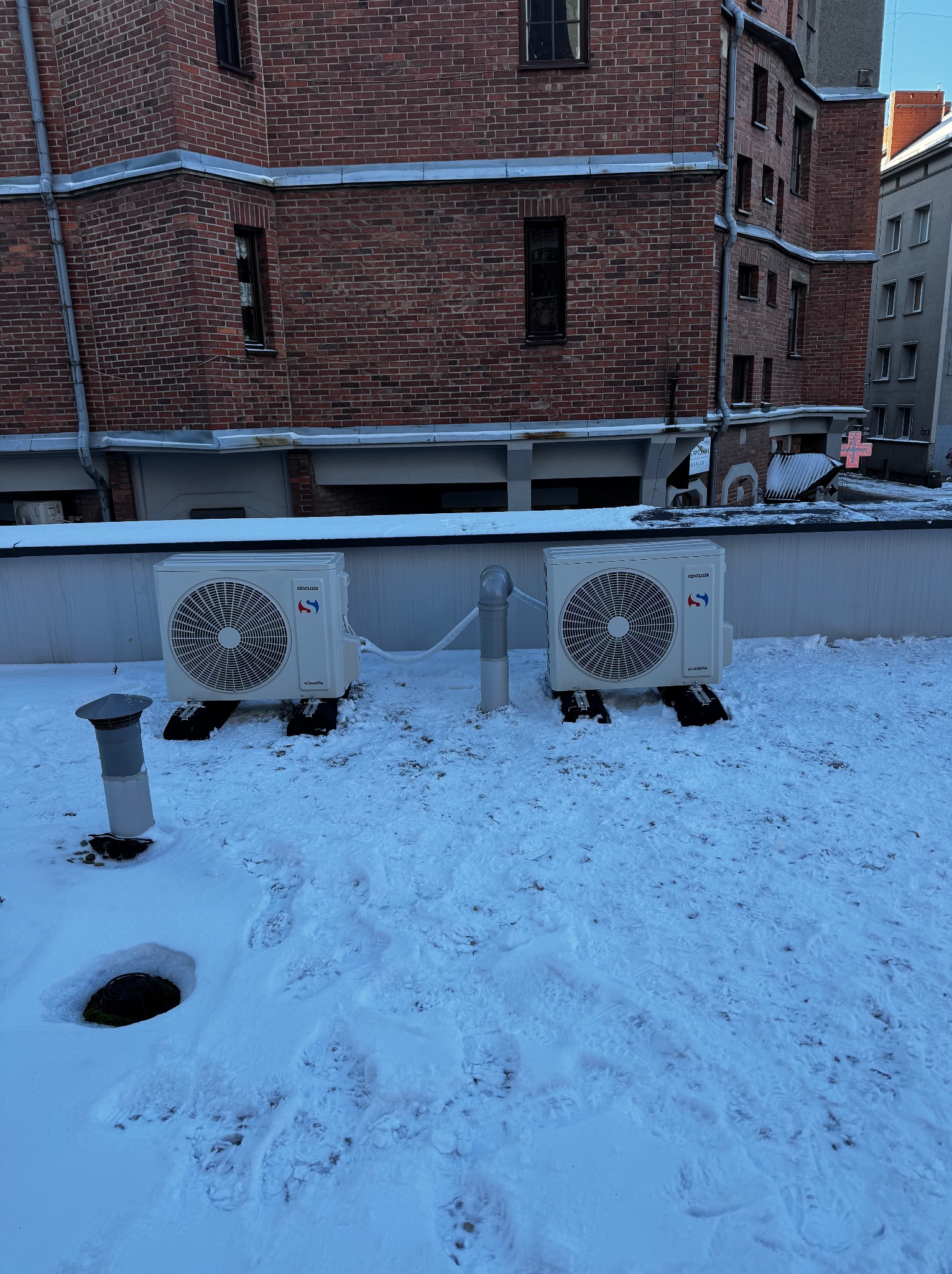 Dwa klimatyzatory Sinclair na dachu budynku pokrytym śniegiem. Widoczne przewody i elementy instalacji. Tło stanowi ceglany budynek i fragment sąsiedniego budynku.