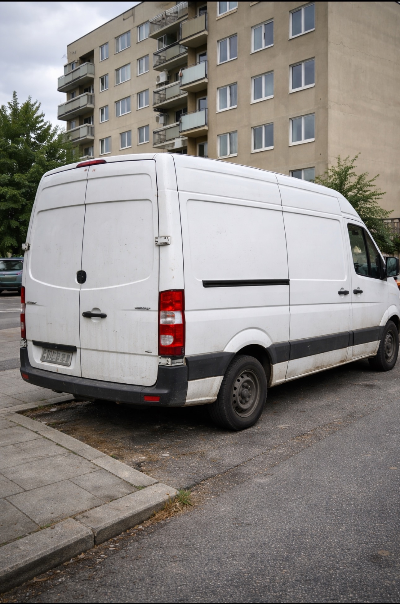 Biały, lekko zabrudzony furgon dostawczy zaparkowany na ulicy, z widocznym budynkiem mieszkalnym w tle. Ujęcie z tyłu pojazdu, zarys opon.