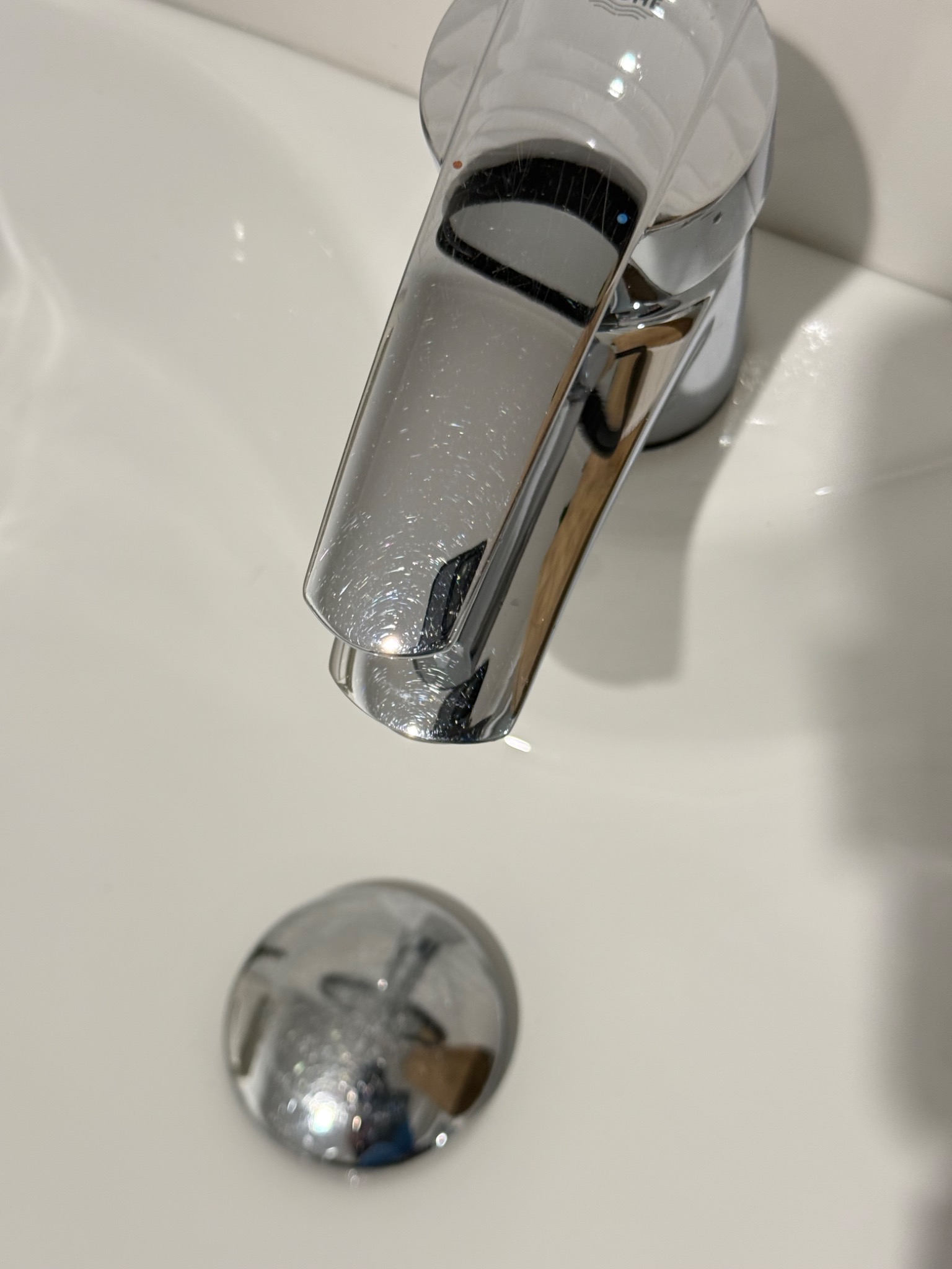 Błyszcząca, chromowana bateria umywalkowa Grohe w białej umywalce. Widoczny korek odpływu. Minimalistyczny design, czystość i połysk.