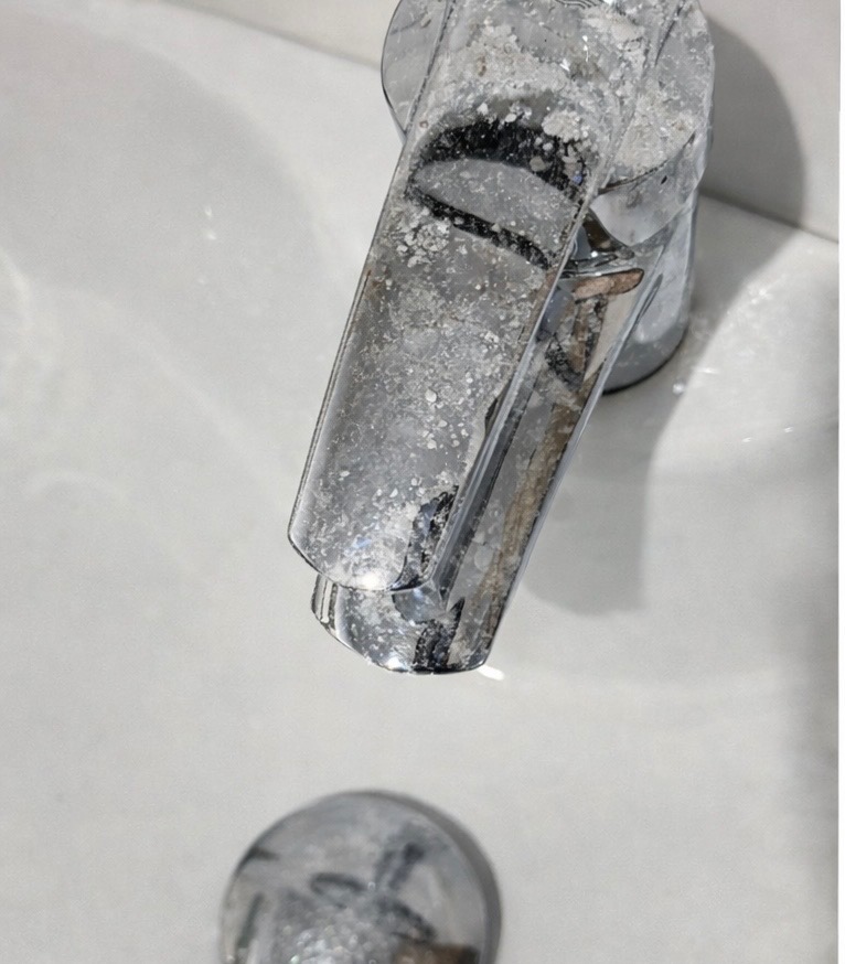 Zakamieniona bateria umywalkowa z bliska, pokryta białym osadem. Widoczny korek umywalki również z osadem. Potrzeba usunięcia kamienia i wyczyszczenia armatury.