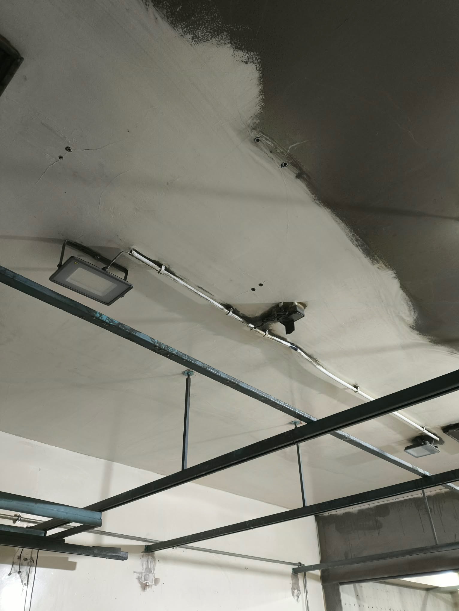 Częściowo pomalowany sufit w pomieszczeniu z widocznymi instalacjami, oświetleniem i metalową konstrukcją. Fragment ściany w tle. Widok z dołu.