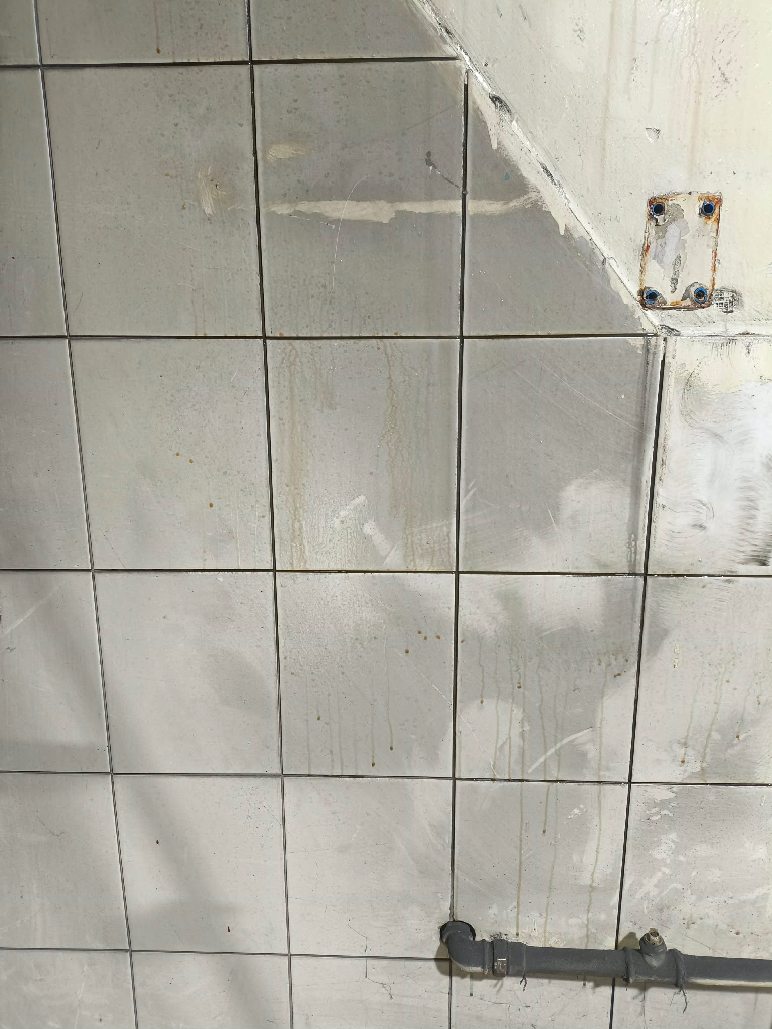 Brudne, białe płytki ścienne z zaciekami i resztkami farby, fragment rury kanalizacyjnej w dolnej części kadru, widoczne ślady po demontażu elementu na ścianie.