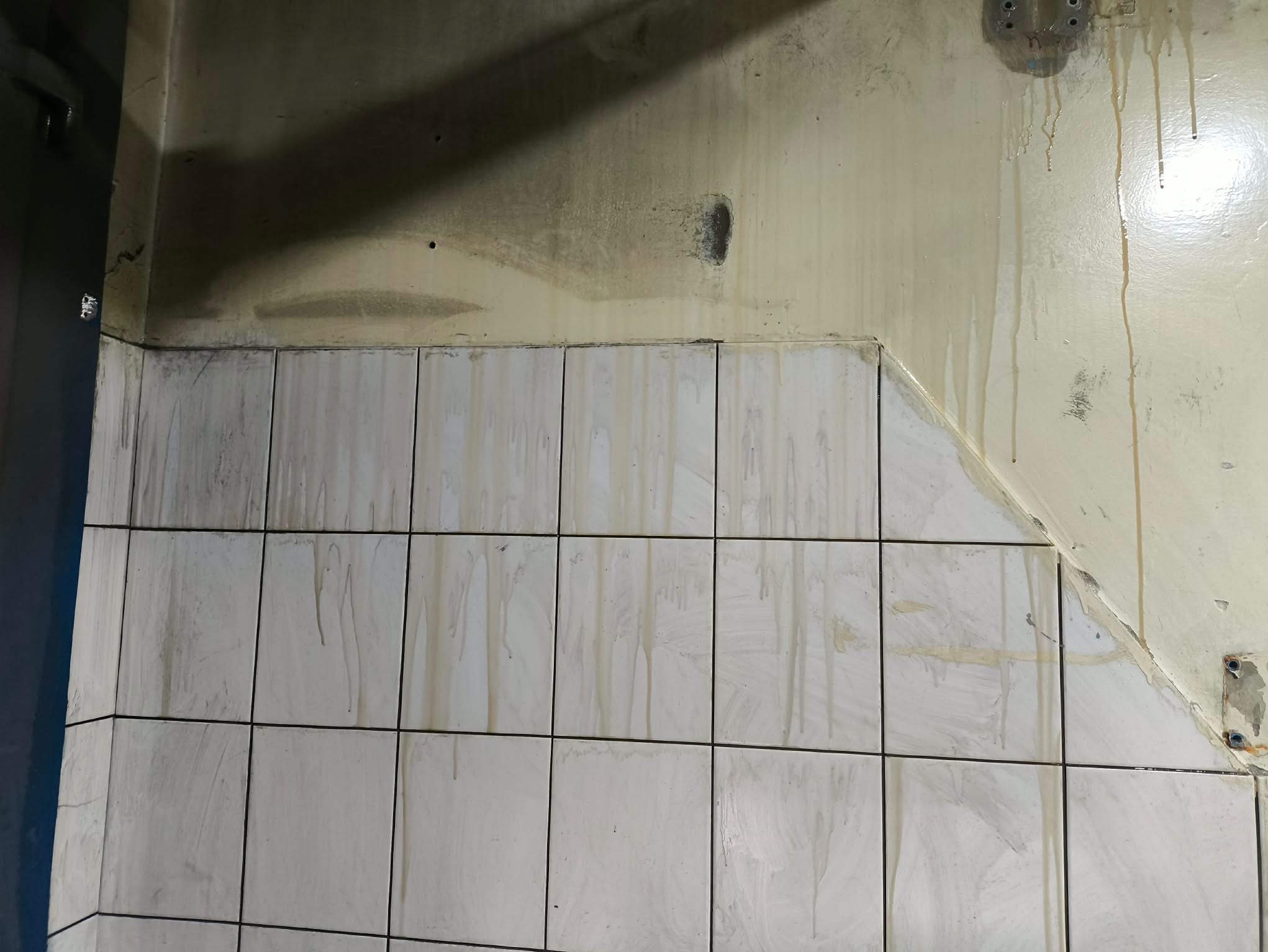 Zabrudzona ściana z białymi płytkami i zaciekami, widoczne ślady po demontażu elementów. Narożnik pomieszczenia z widocznymi plamami i zabrudzeniami na ścianie.